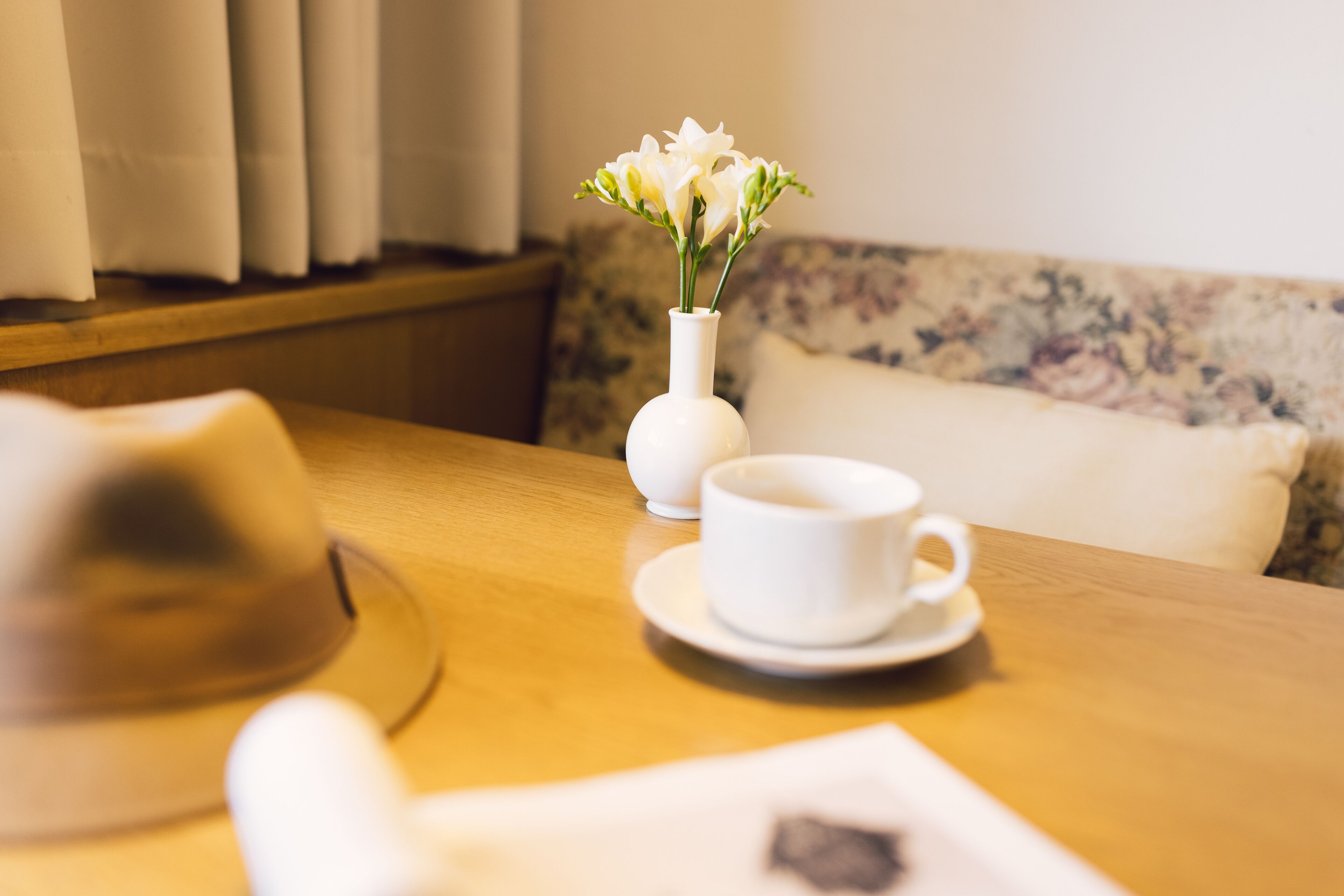 Una mesa de madera con una taza y platillo blancos, un jarrón blanco con flores y un objeto desenfocado al frente.