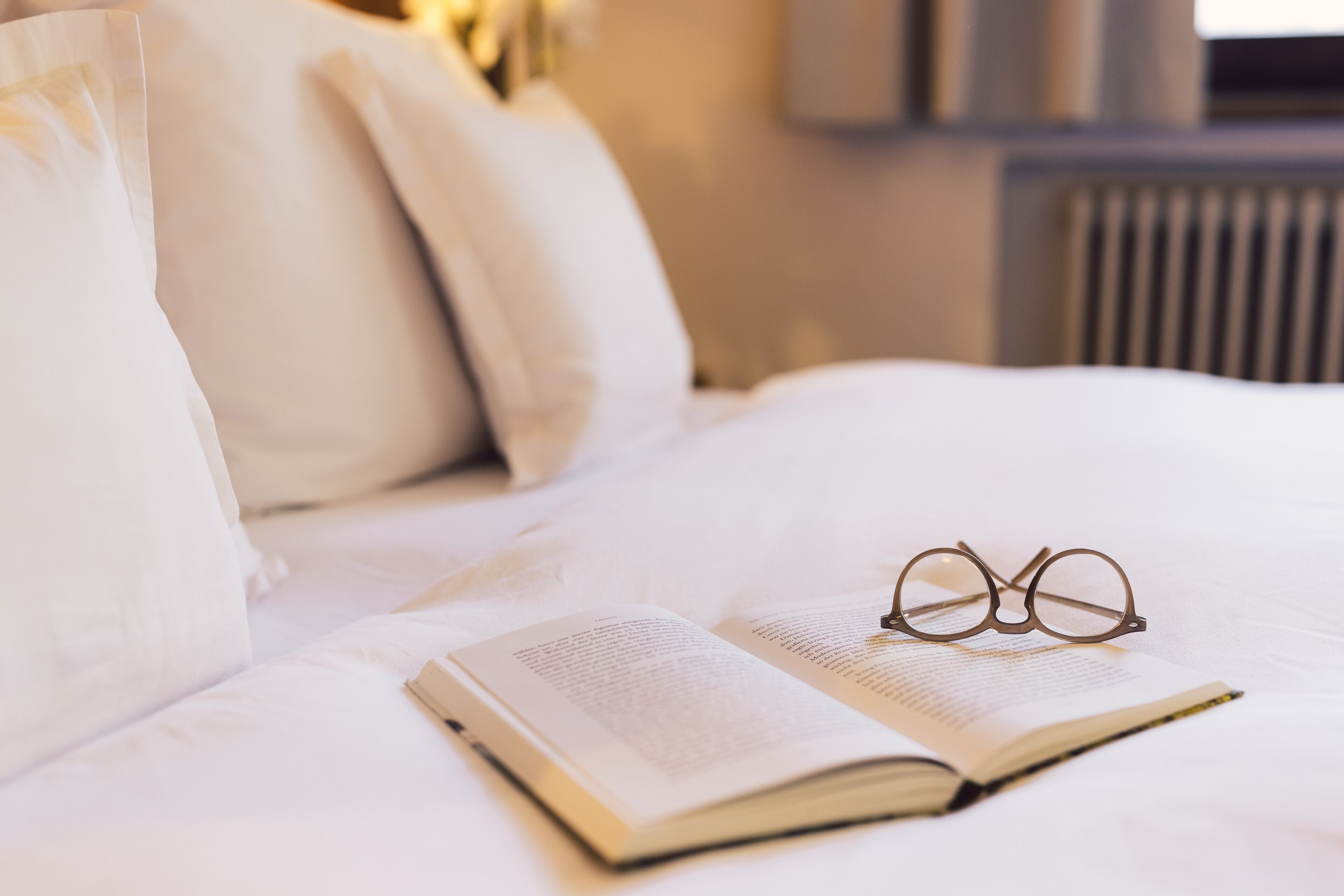 A neatly made bed with white linens, a book, and a pair of glasses on it.