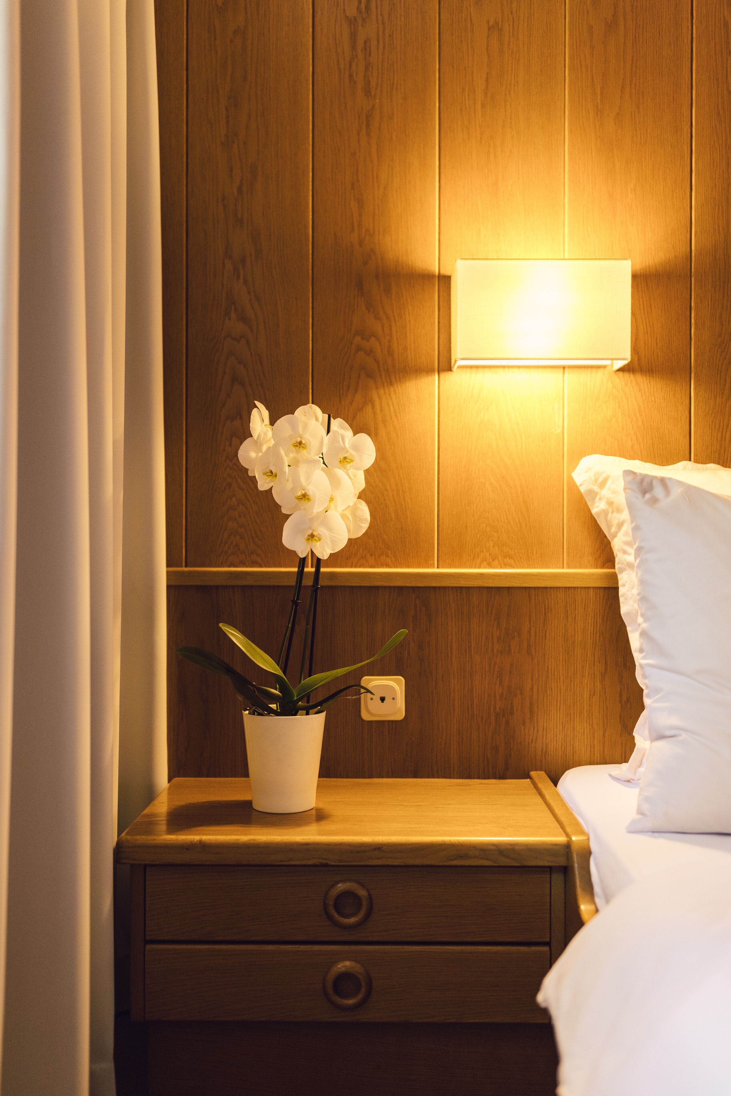 Une table de chevet en bois, sur laquelle se trouve une orchidée blanche dans un pot, une lampe de chevet et un oreiller blanc sur un lit.