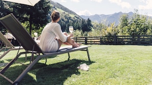 Una persona sentada en una silla de descanso bajo una sombrilla, sosteniendo un vaso de bebida, con vistas a montañas y árboles en el fondo.