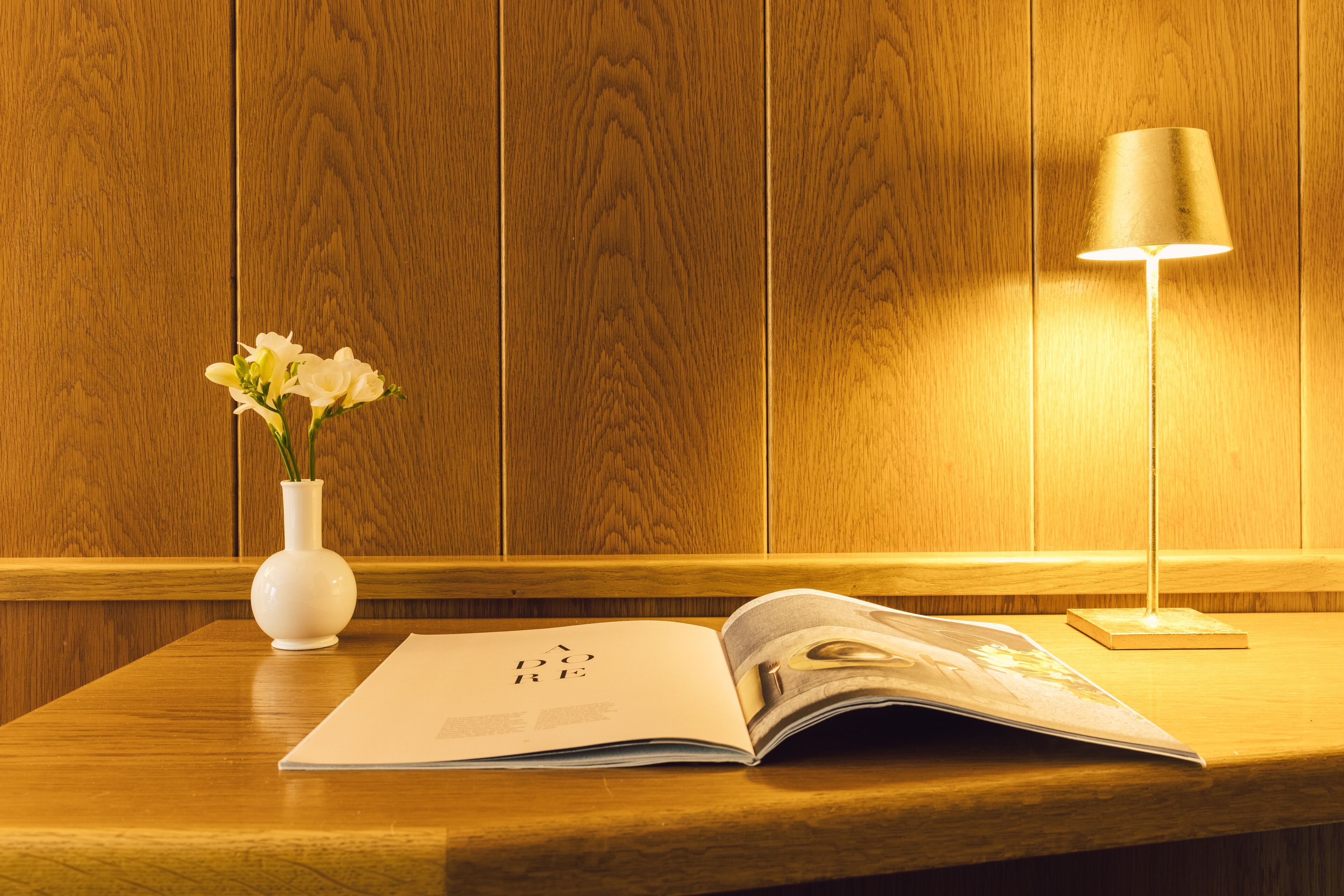 A wooden table with a white vase containing flowers, an open magazine, and a lamp.