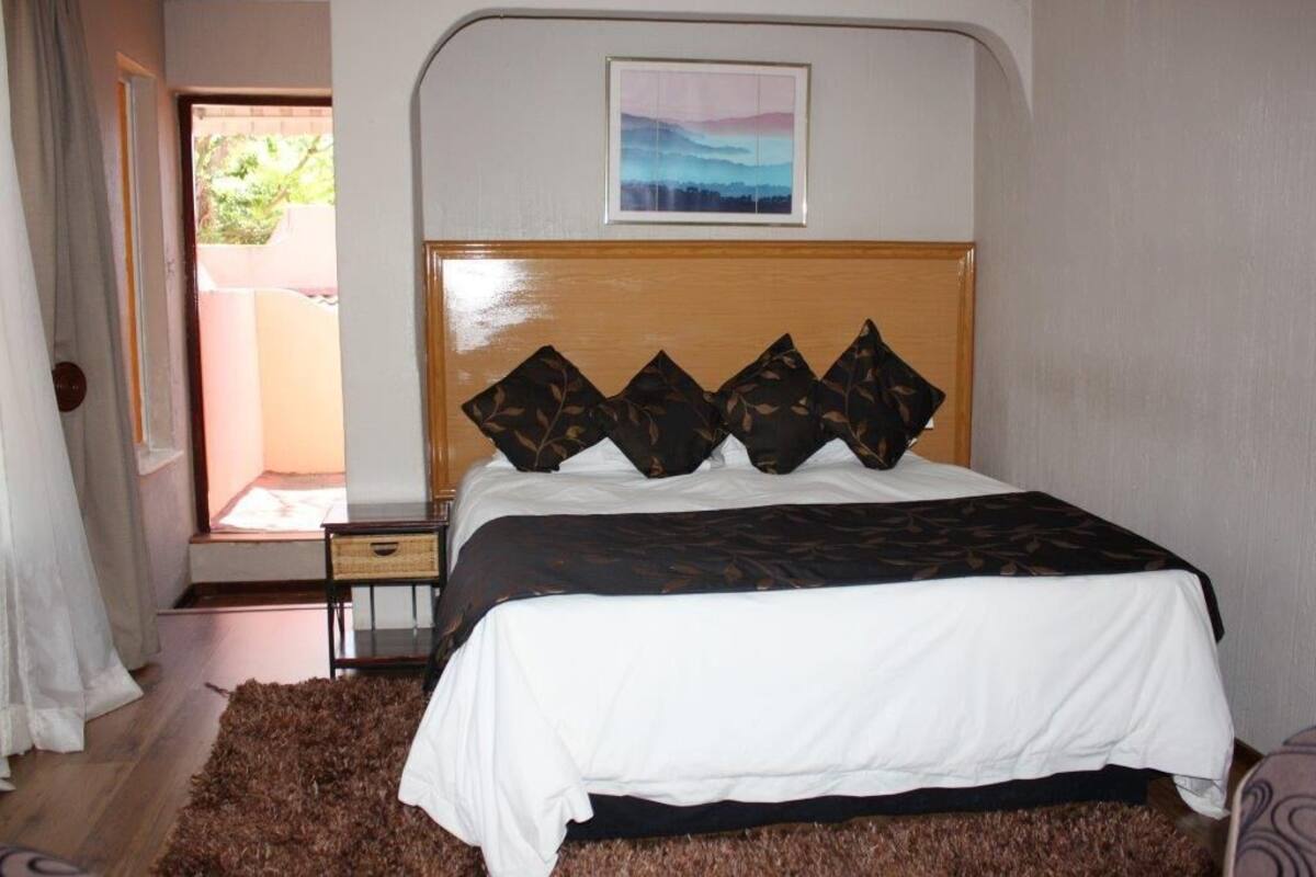 A bedroom with a bed, a wooden headboard, a framed picture above the bed, and a view of the outside through an open door.