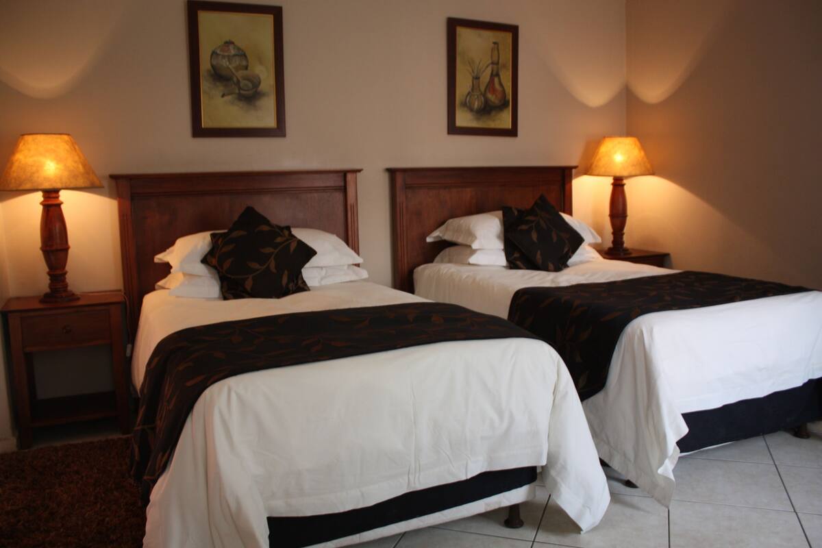 A hotel room with two beds, each with a wooden headboard, white bedding, and a dark-colored throw blanket. There are two bedside lamps on wooden nightstands.