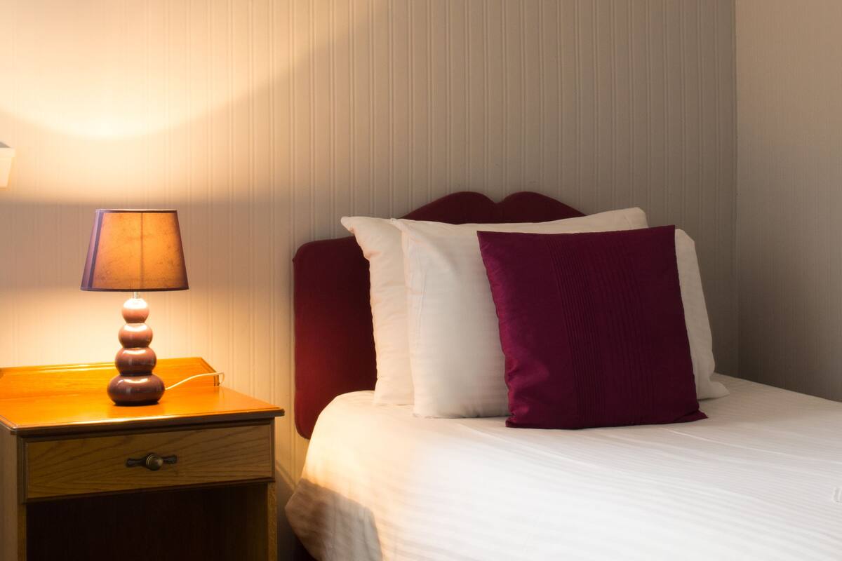 A neatly made bed with white and purple pillows, a wooden nightstand with a lamp, and a closed drawer.