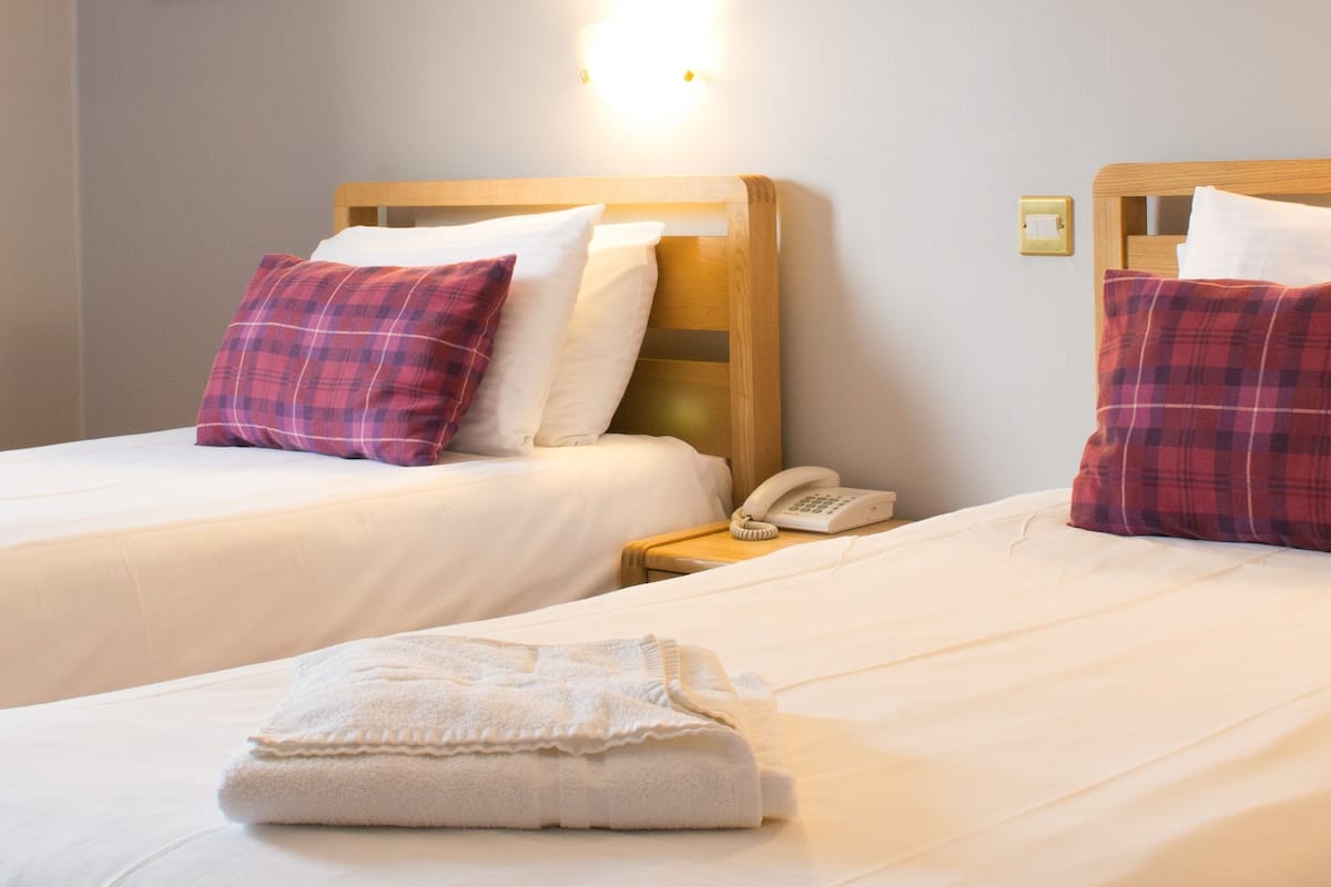A hotel room with two beds, each with a wooden headboard, white bedding, and red plaid pillows. There is a bedside table with a telephone and a light switch on the wall.