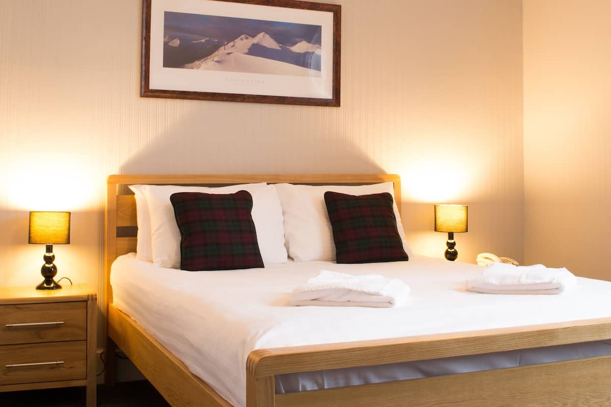 A neatly made bed with white linens and plaid pillows, flanked by two bedside lamps. A framed photograph of a mountain landscape hangs above the bed.