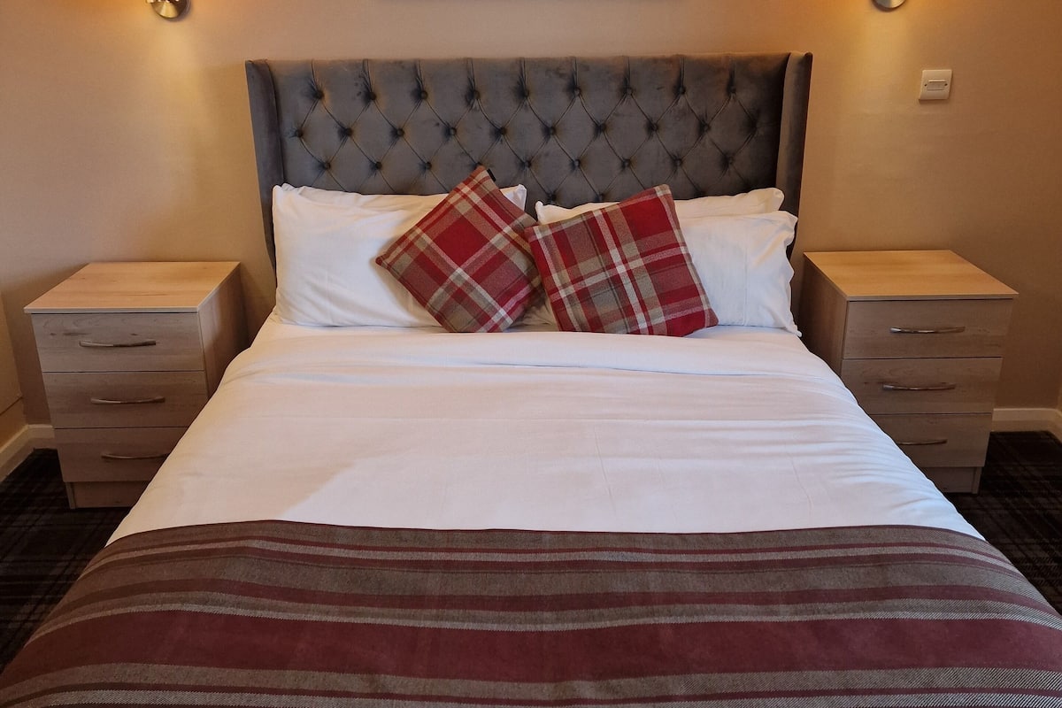 A neatly made bed with a striped blanket, two plaid pillows, and a tufted headboard. There are two bedside tables with lamps and a painting of a mountain scene above the bed.