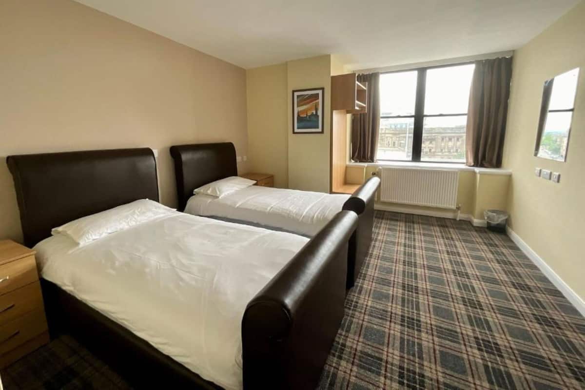 A hotel room with two beds, a window with curtains, a radiator, and a plaid carpet.