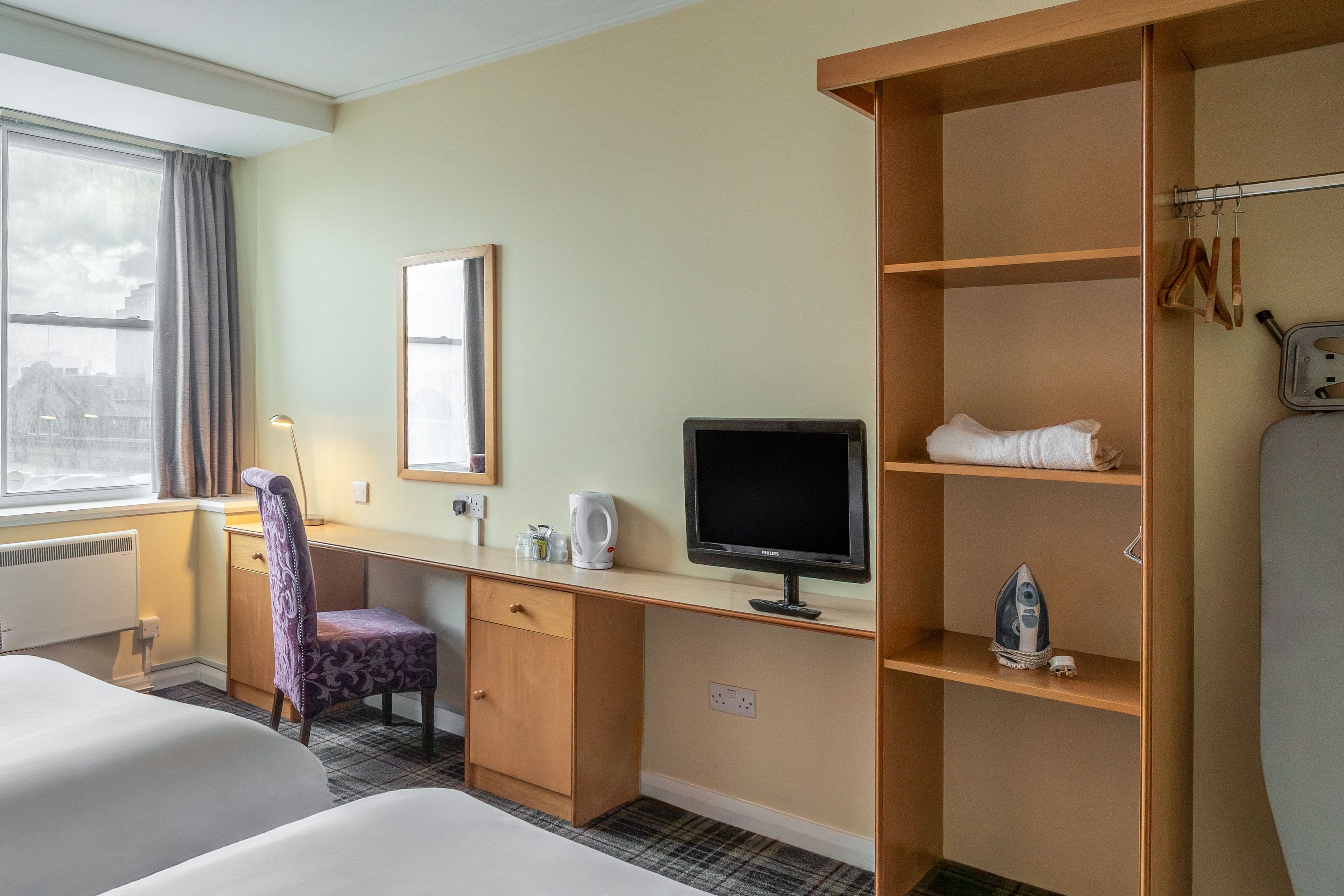 A hotel room with a bed, a desk with a computer, a television, and a shelf with towels and a kettle.
