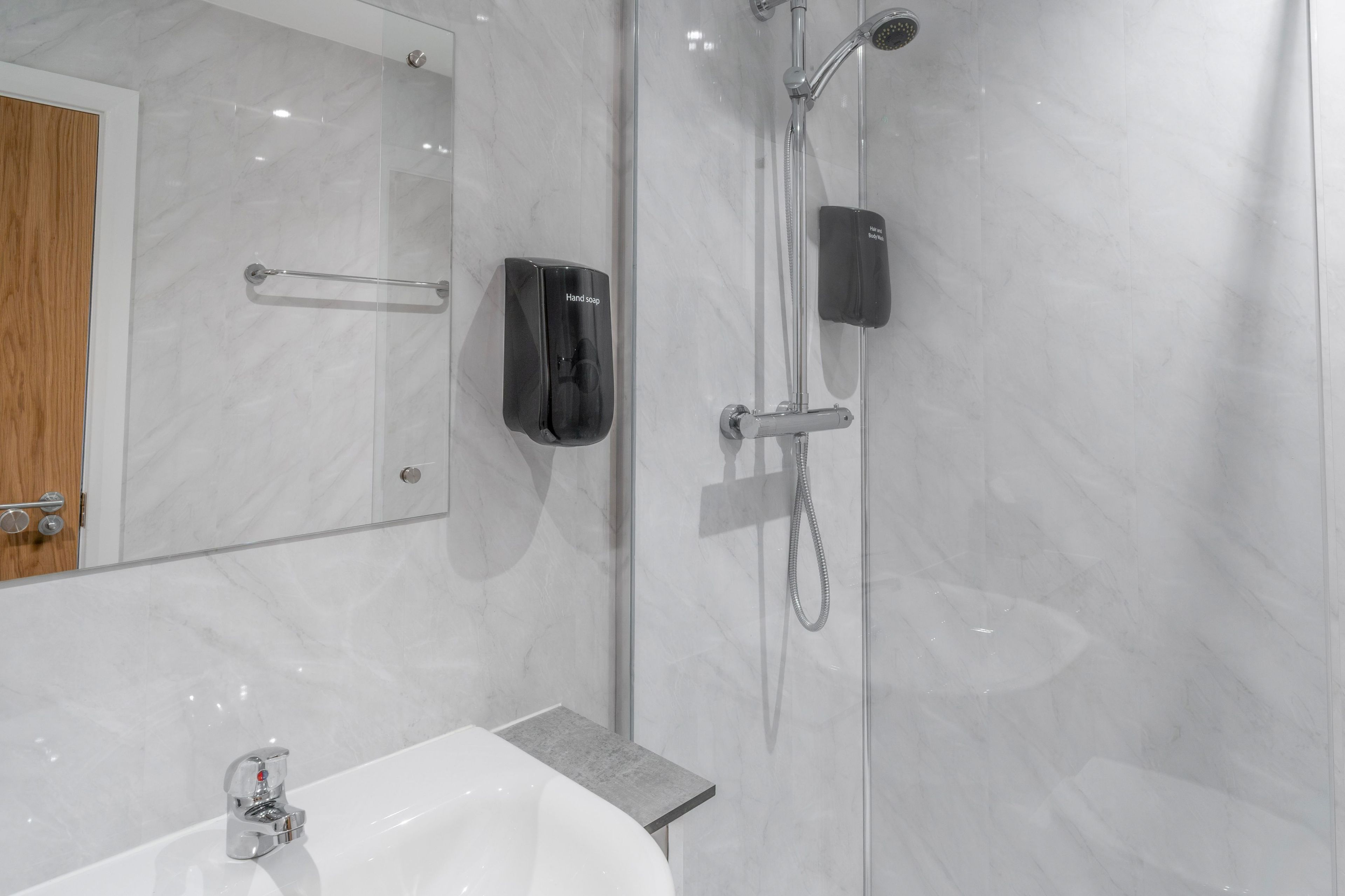 A modern bathroom with a marble wall, a hand soap dispenser, a handheld showerhead, and a sink with a chrome faucet.