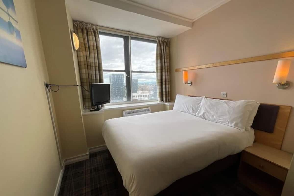A hotel room with a large bed, a window with curtains, a flat-screen TV, and two wall-mounted lights.