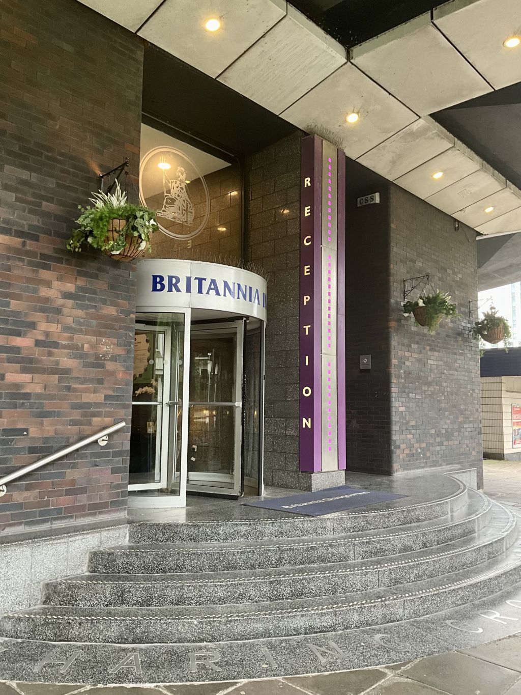 Un espace d'accueil avec une entrée dotée d'une porte en verre, une enseigne indiquant « BRITANNIA », et un escalier menant à l'entrée.