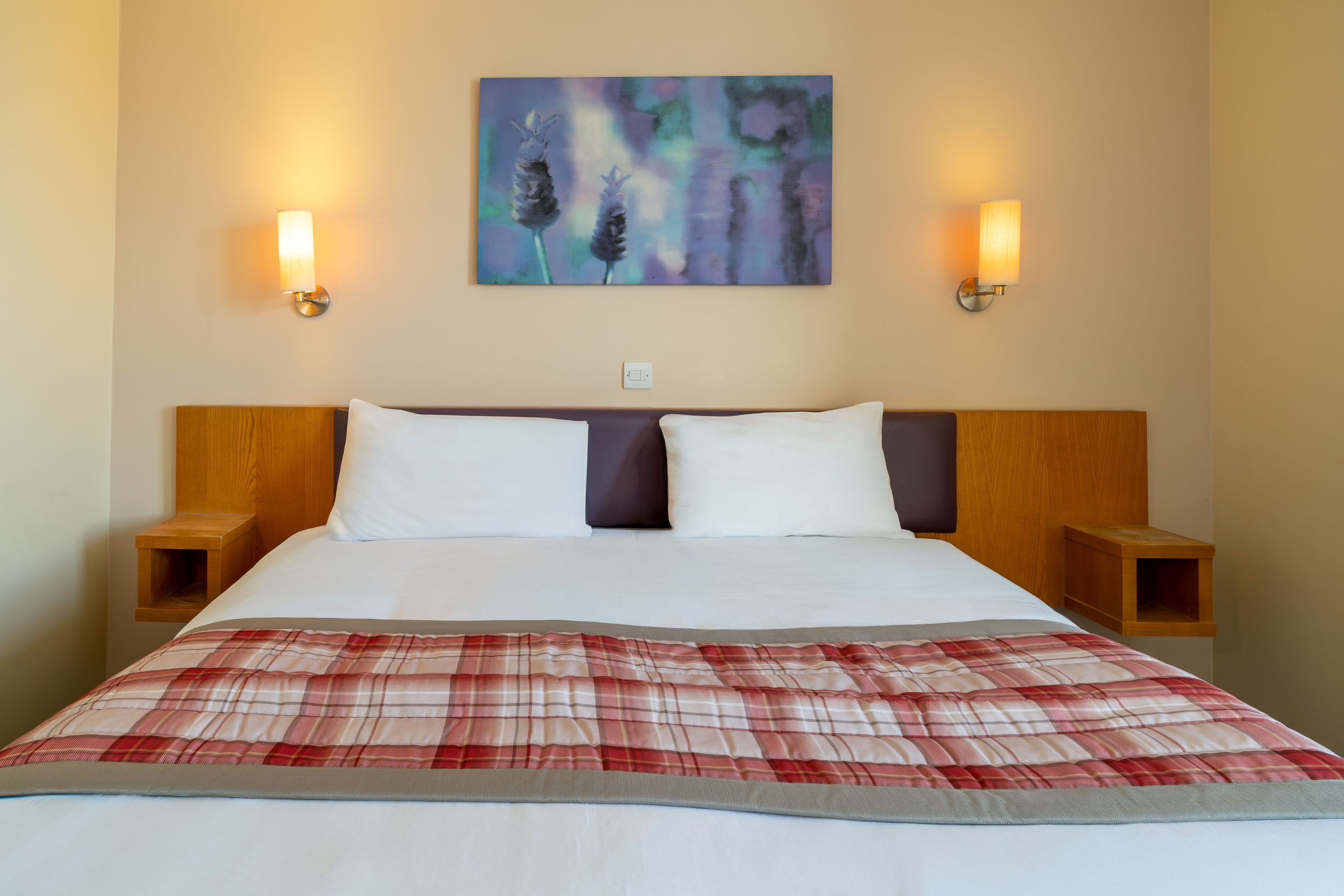 Ein Hotelzimmer mit einem Bett, zwei Nachttischen, einem Kopfteil, einem Bild an der Wand und zwei Wandleuchten.