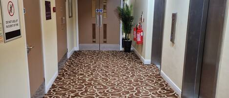 Un pasillo de hotel con alfombra estampada, puertas dobles, extintores y un ascensor.