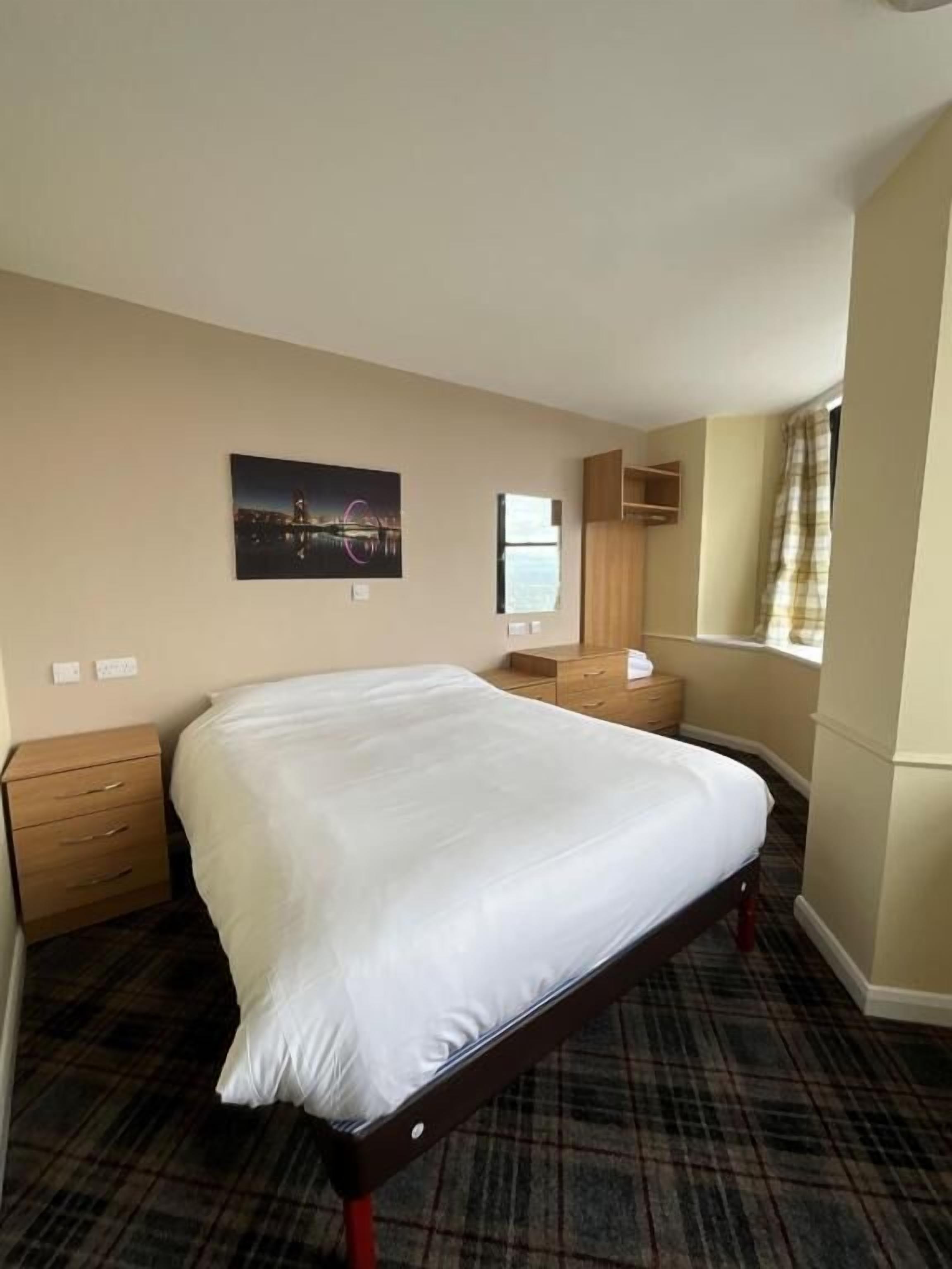 Una habitación de hotel con una cama grande, una mesita de noche de madera, un tocador pequeño, una ventana con vistas y un cuadro en la pared.
