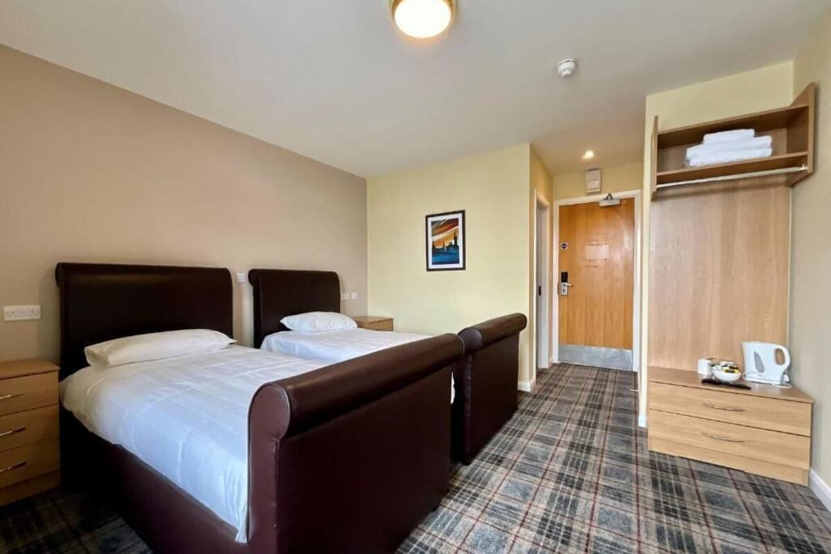 A hotel room with two beds, a wooden wardrobe, a plaid carpet, and a framed picture on the wall.