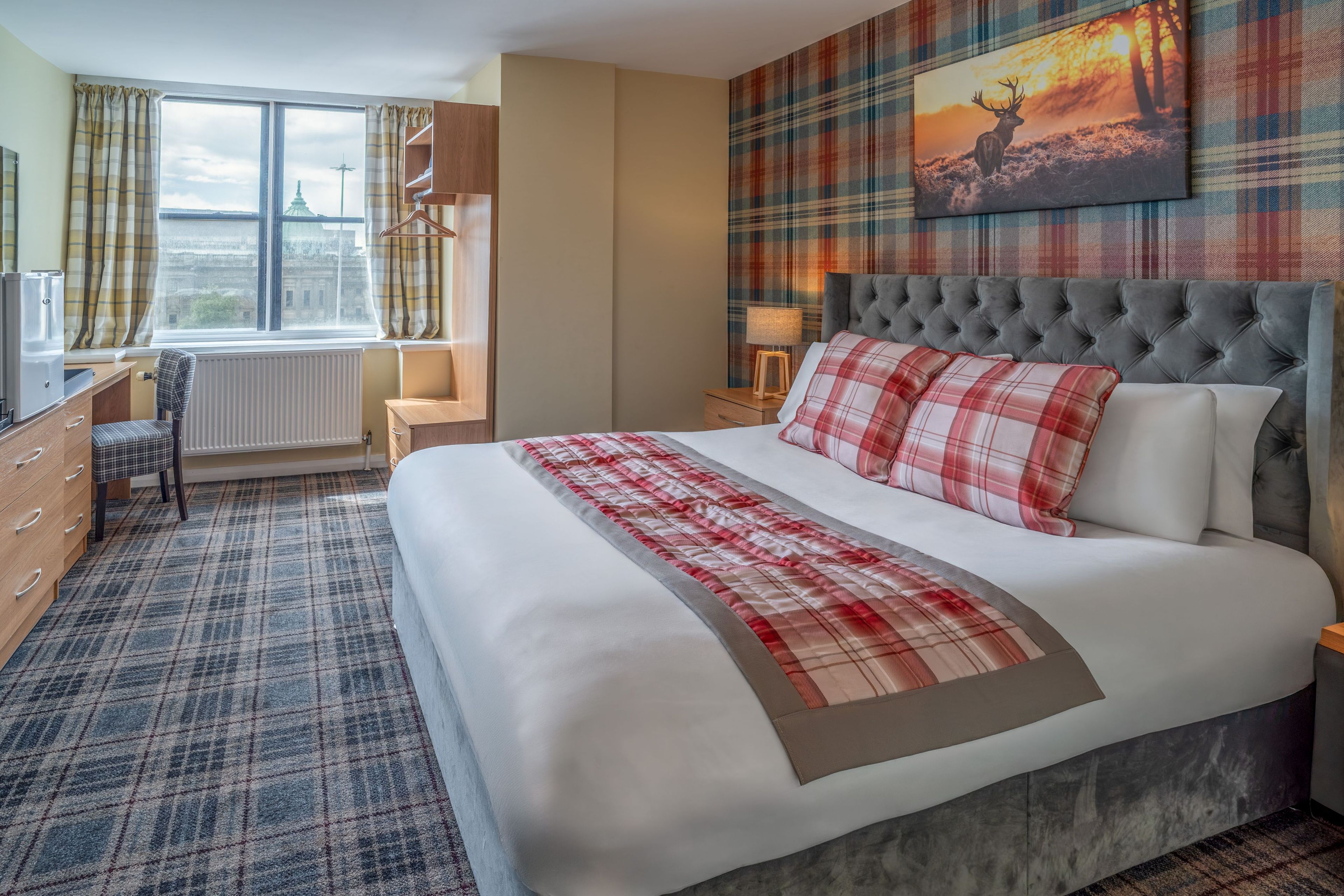 A hotel room with a bed, a desk, a chair, a radiator, a window with curtains, and a painting on the wall.