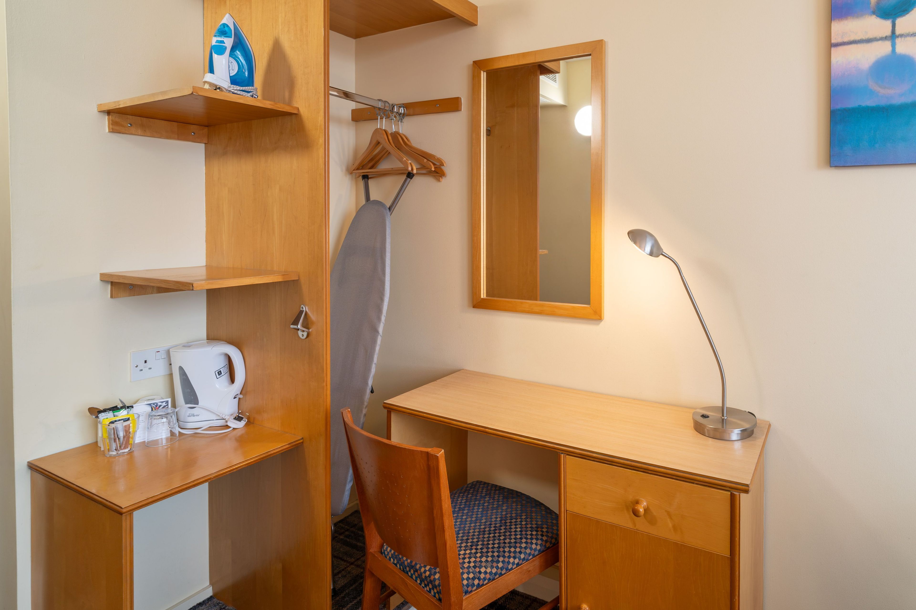 A hotel room with a wooden desk, a chair, a mirror, a hanging clothes rack, and a kettle.