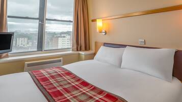 A hotel room with a bed, a window with curtains, a wall-mounted light, and a view of buildings outside.