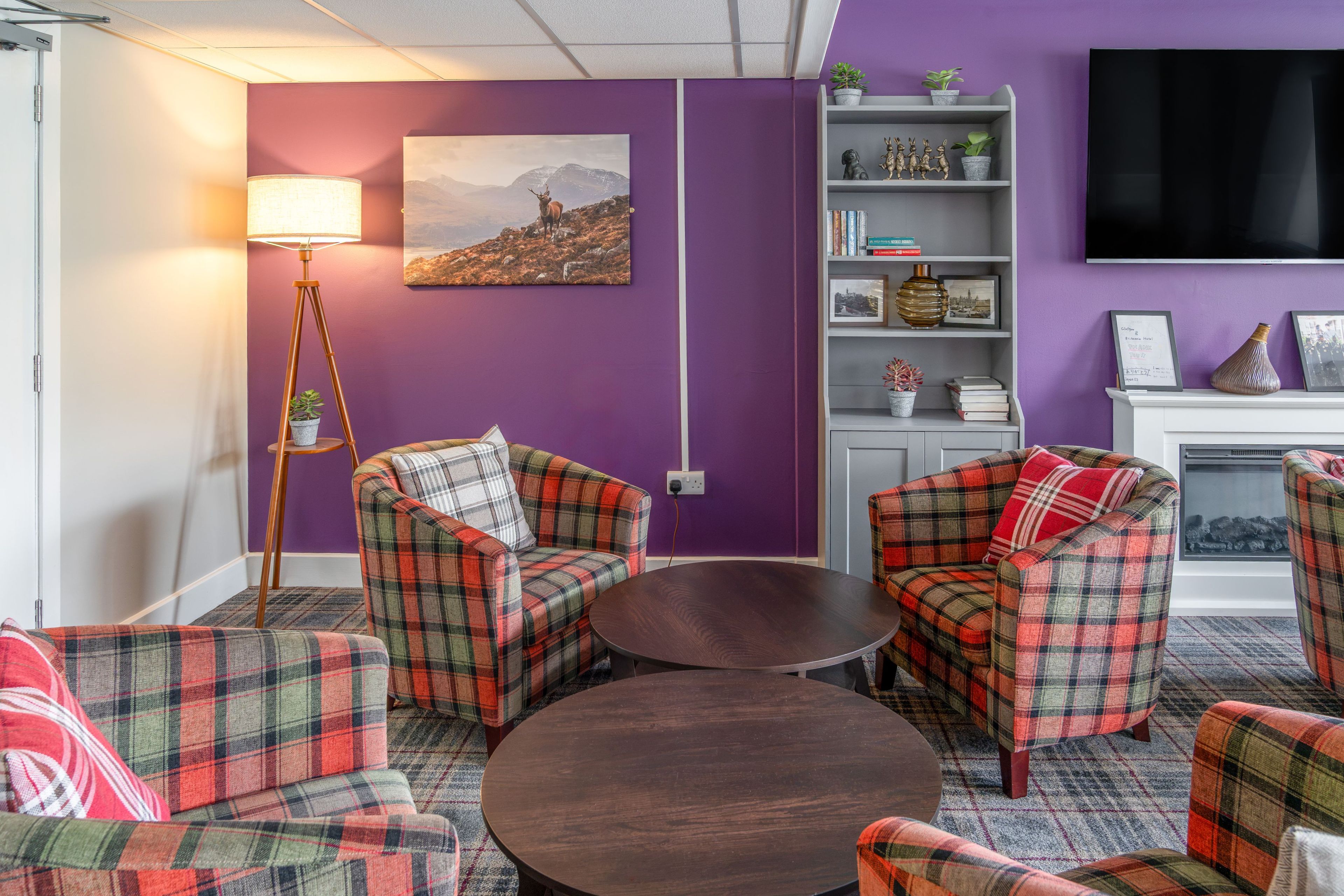 A room with purple walls, plaid furniture, a flat-screen TV, a bookshelf, and a painting.