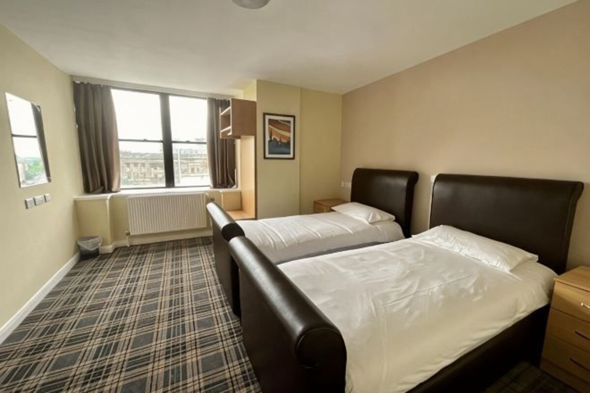 A hotel room with two beds, a window with curtains, a radiator, and a picture on the wall.