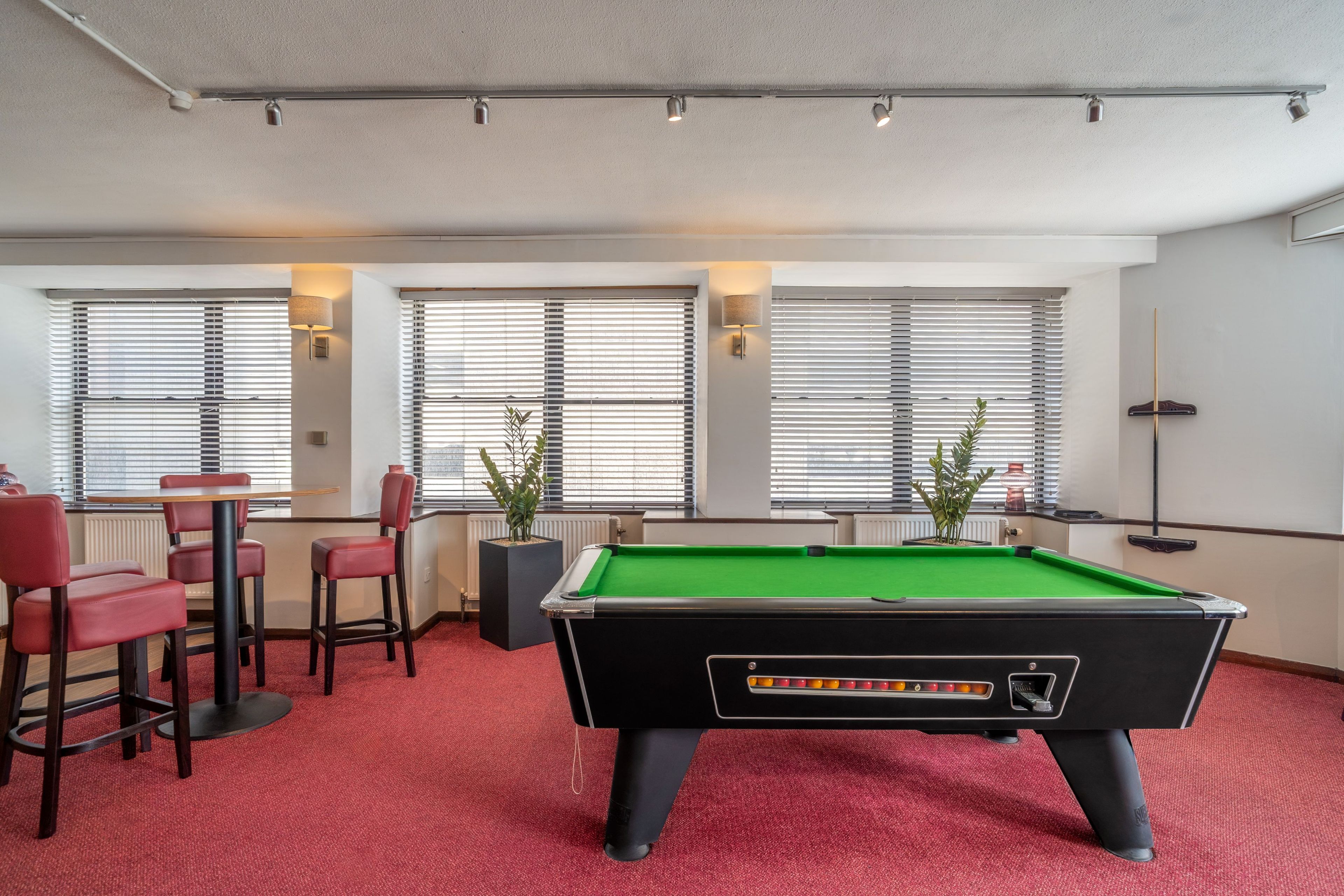 A pool table in a room with red carpet, a bar area with stools, and large windows with blinds.