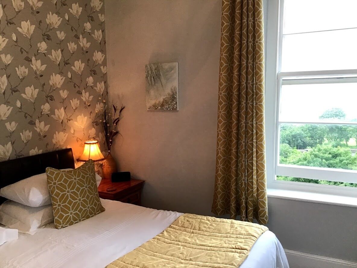Een opgemaakt bed met een wit-geel sprei, een bedlamp en een ingelijst foto aan de muur.