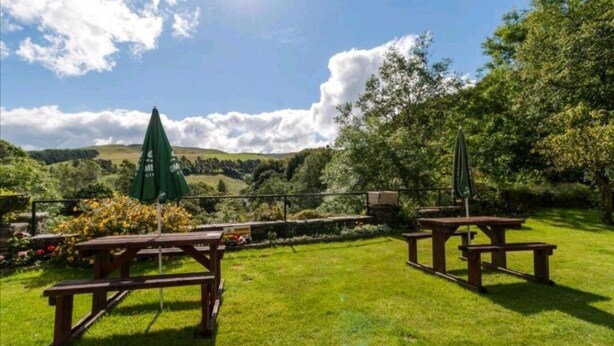 A picnic area with wooden benches and a green umbrella, surrounded by lush greenery and hills in the background. - George Borrow Hotel (Aberystwyth)