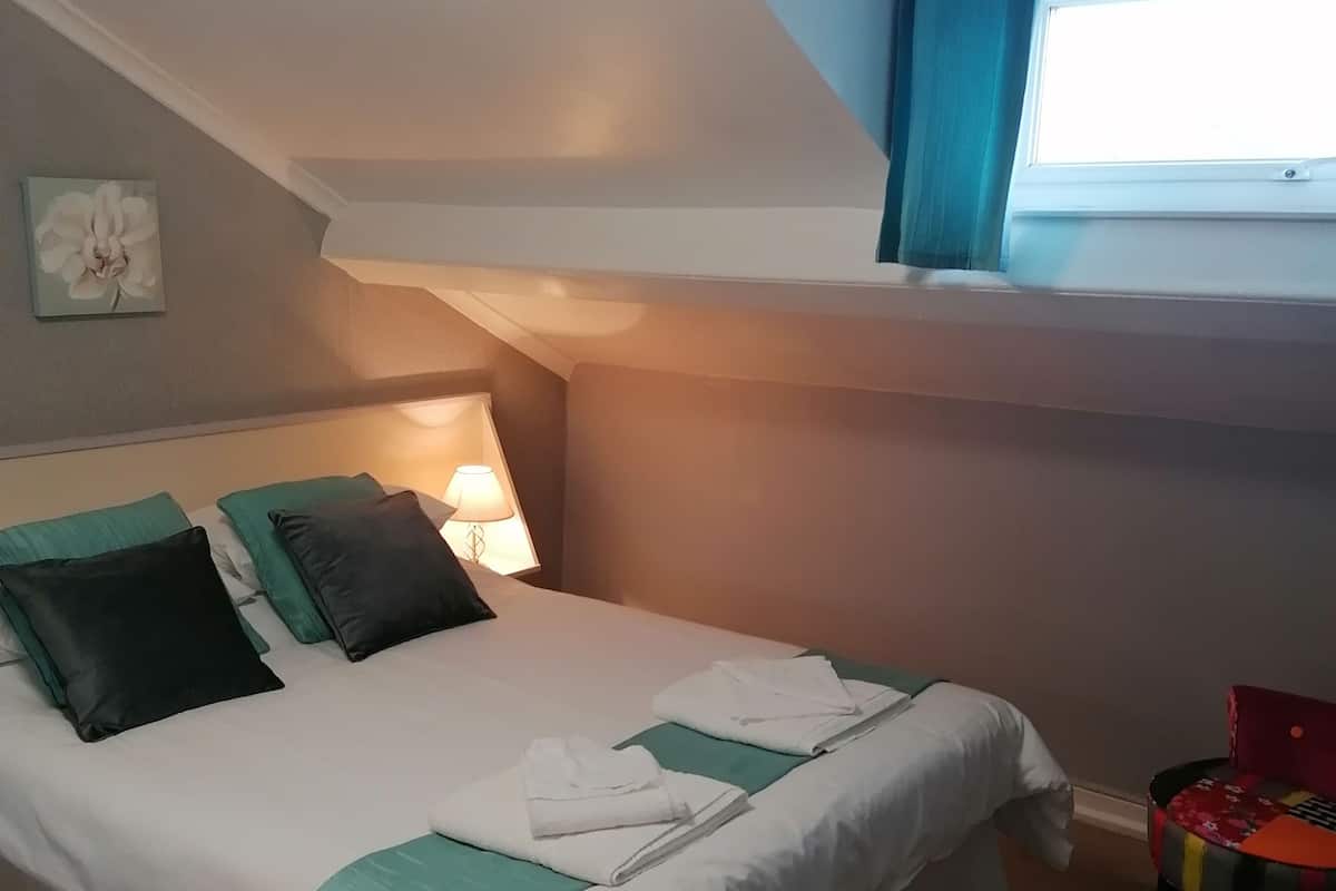 A neatly made bed with white and teal bedding, a single green pillow, and a black pillow. There is a small table with a lamp beside the bed. A chair with a colorful cushion is visible in the corner.
