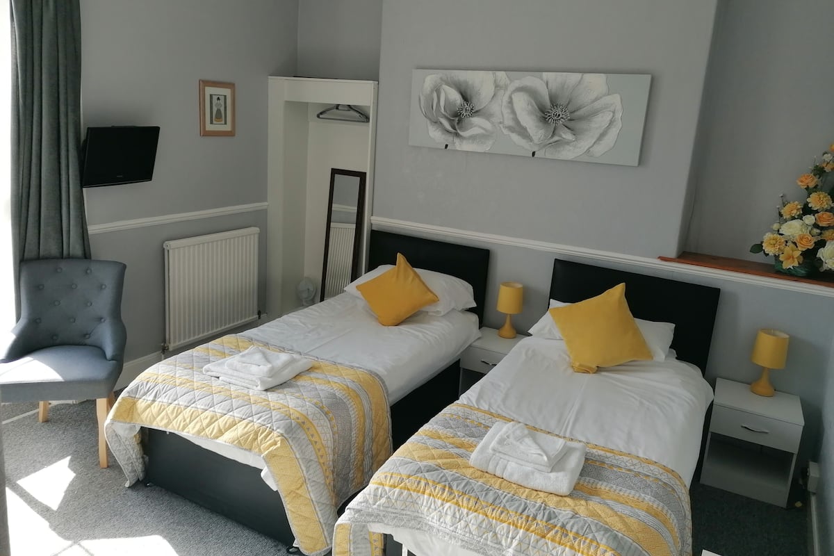 A hotel room with two beds, each with a white and yellow patterned bedspread, a mirror, a flat-screen TV, and a flower arrangement on the dresser.