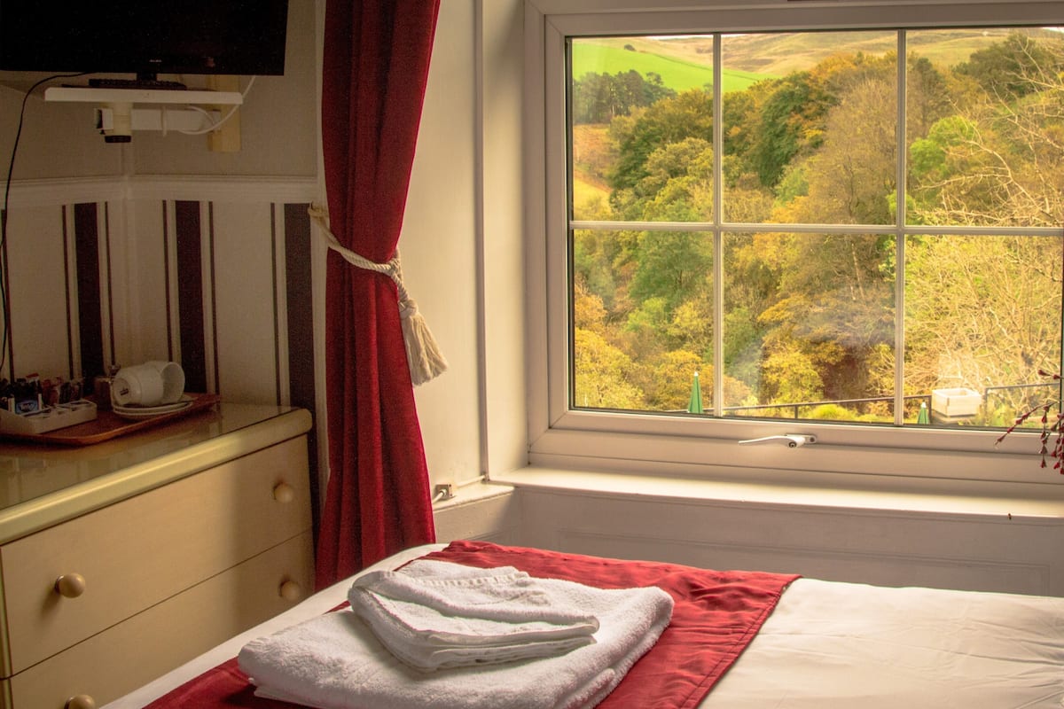 A hotel room with a bed, a window with a view of a forest, a red towel on the bed, and a small table with toiletries.