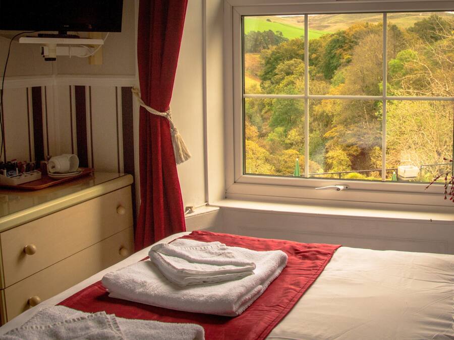 Una habitación de hotel con cama, ventana con vista a un bosque, una toalla roja sobre la cama y una mesita con artículos de tocador.