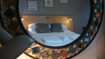 A bed with pillows and a folded towel, a bedside table with a lamp, and a wall-mounted mirror with a decorative frame.