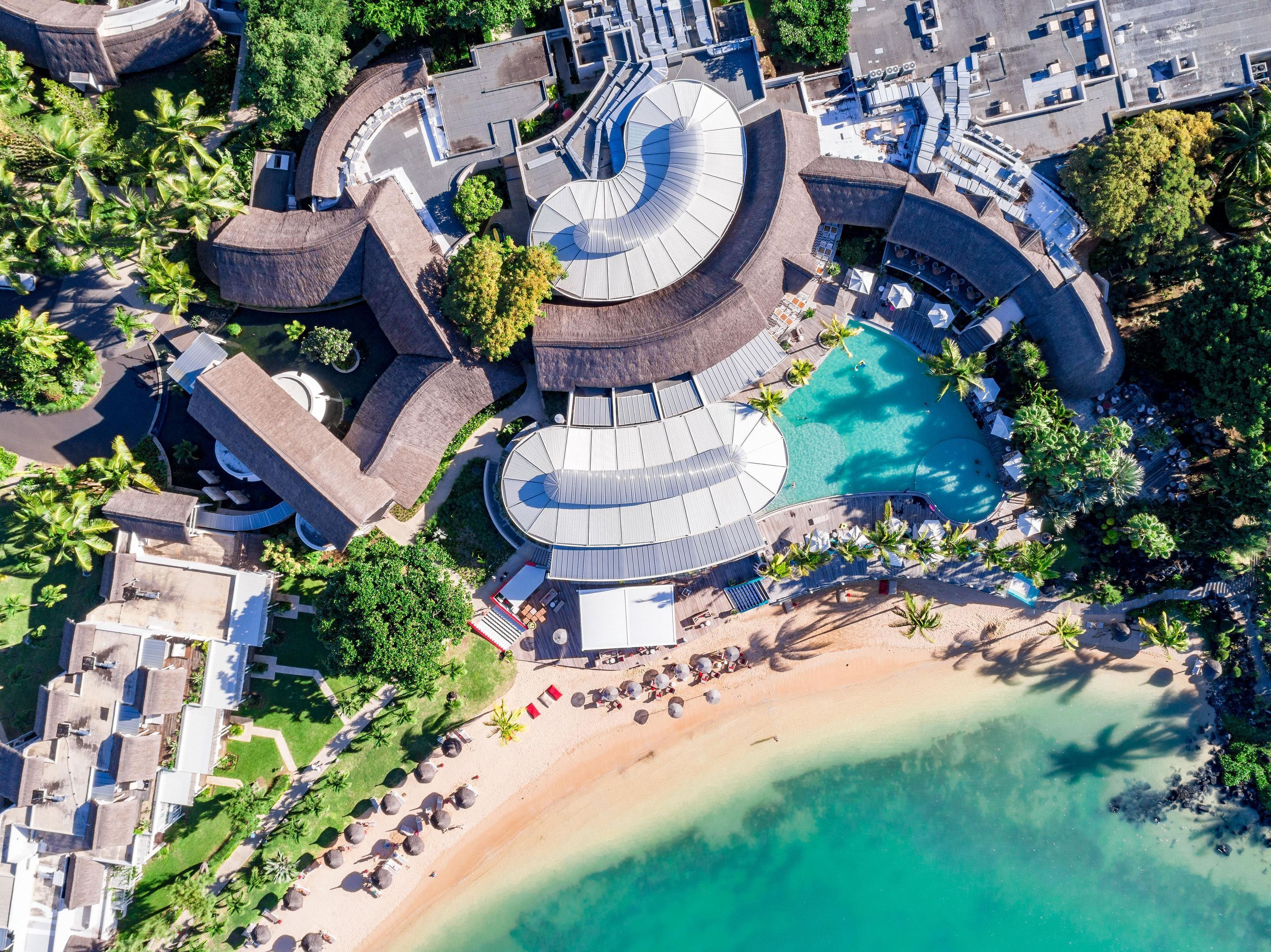 Vue aérienne d’un complexe hôtelier comprenant une piscine centrale, plusieurs bâtiments et une zone de plage.