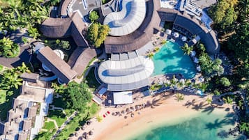 Vue aérienne d’un complexe hôtelier comprenant une piscine centrale, plusieurs bâtiments et une zone de plage.