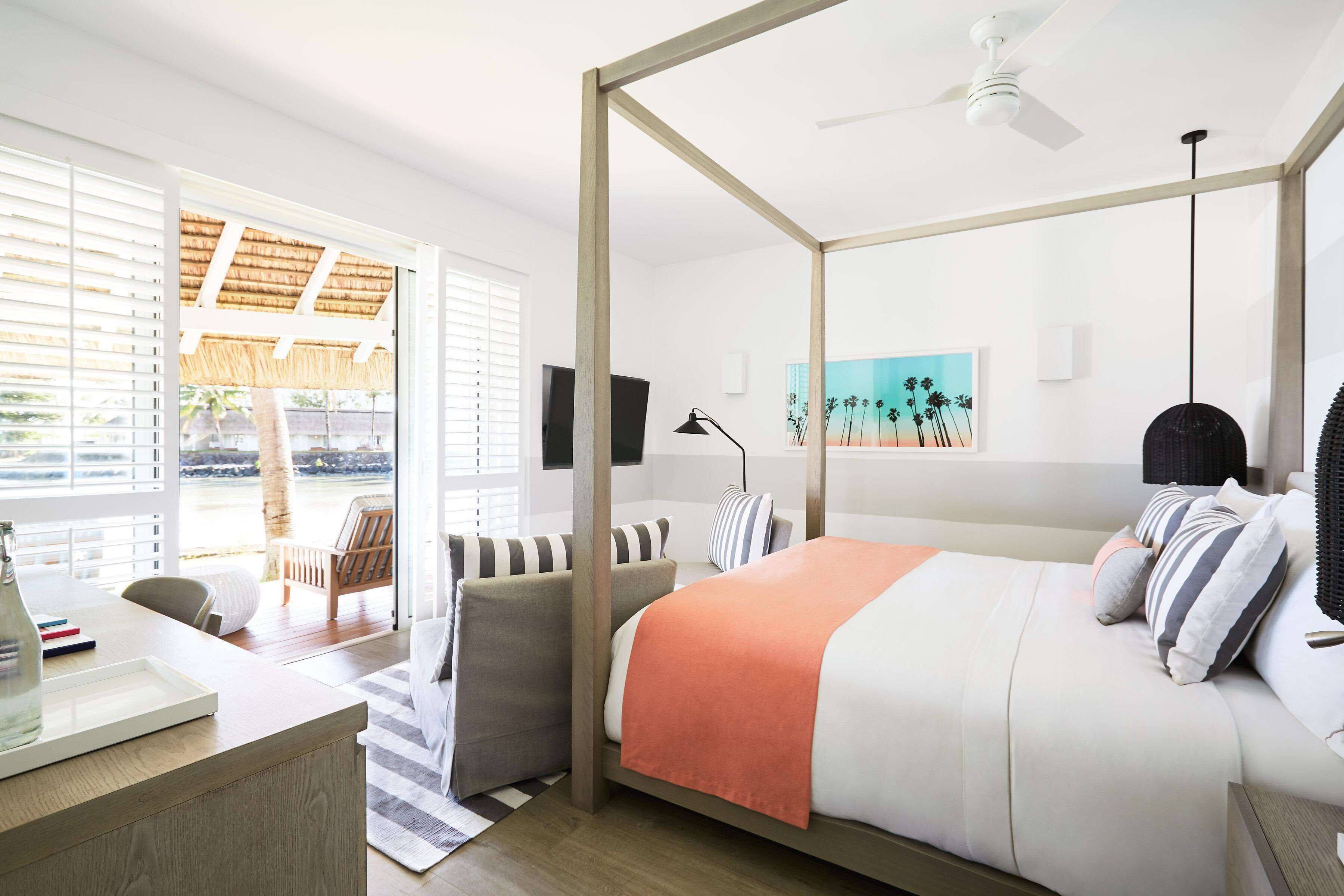 A four-poster bed with a white and orange bedspread, a wooden desk with a tray, a chair, and a view of the outdoors through a glass door.