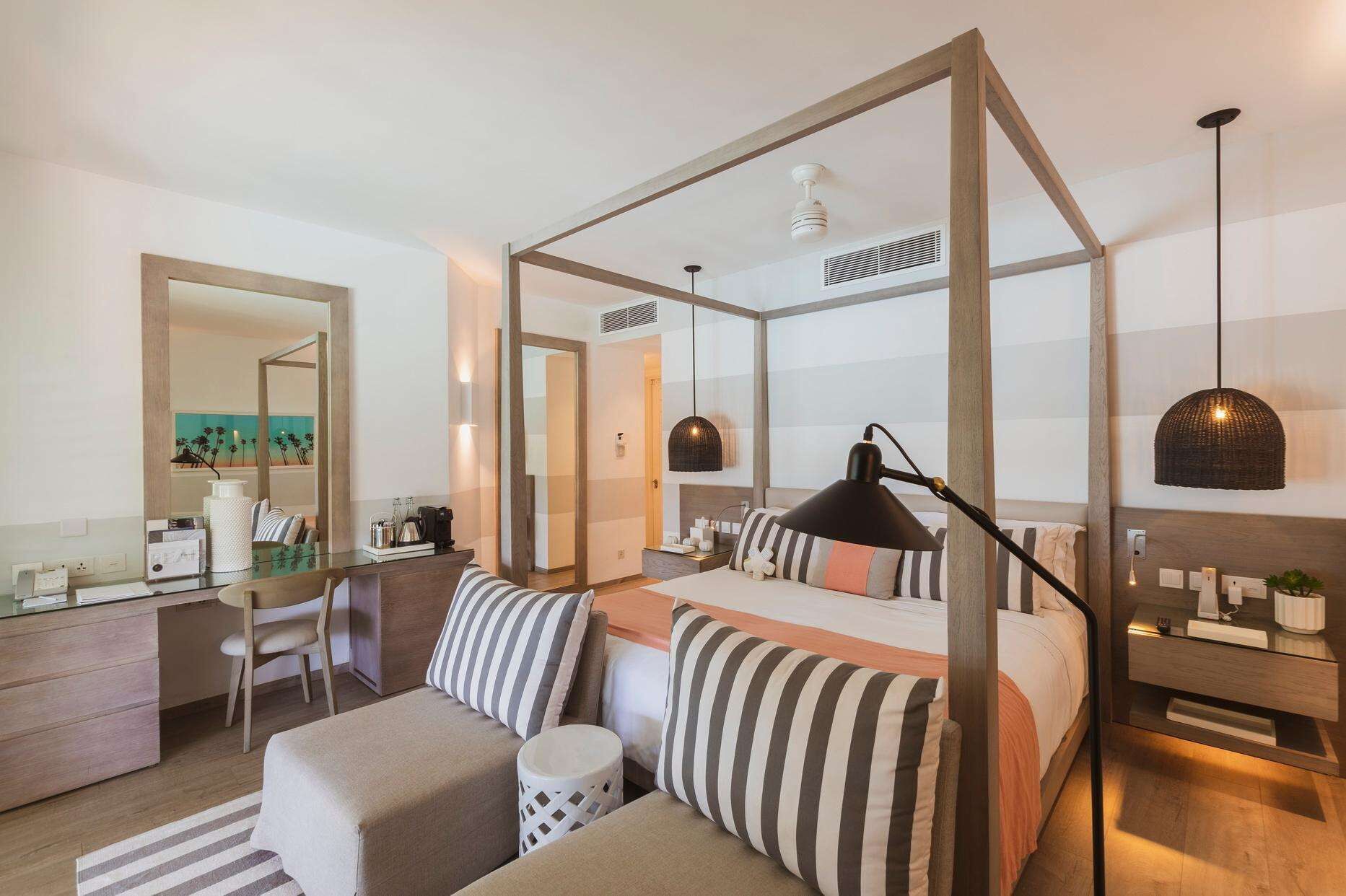 A modern hotel room with a four-poster bed, a desk with a chair, a small round table, and a large mirror.