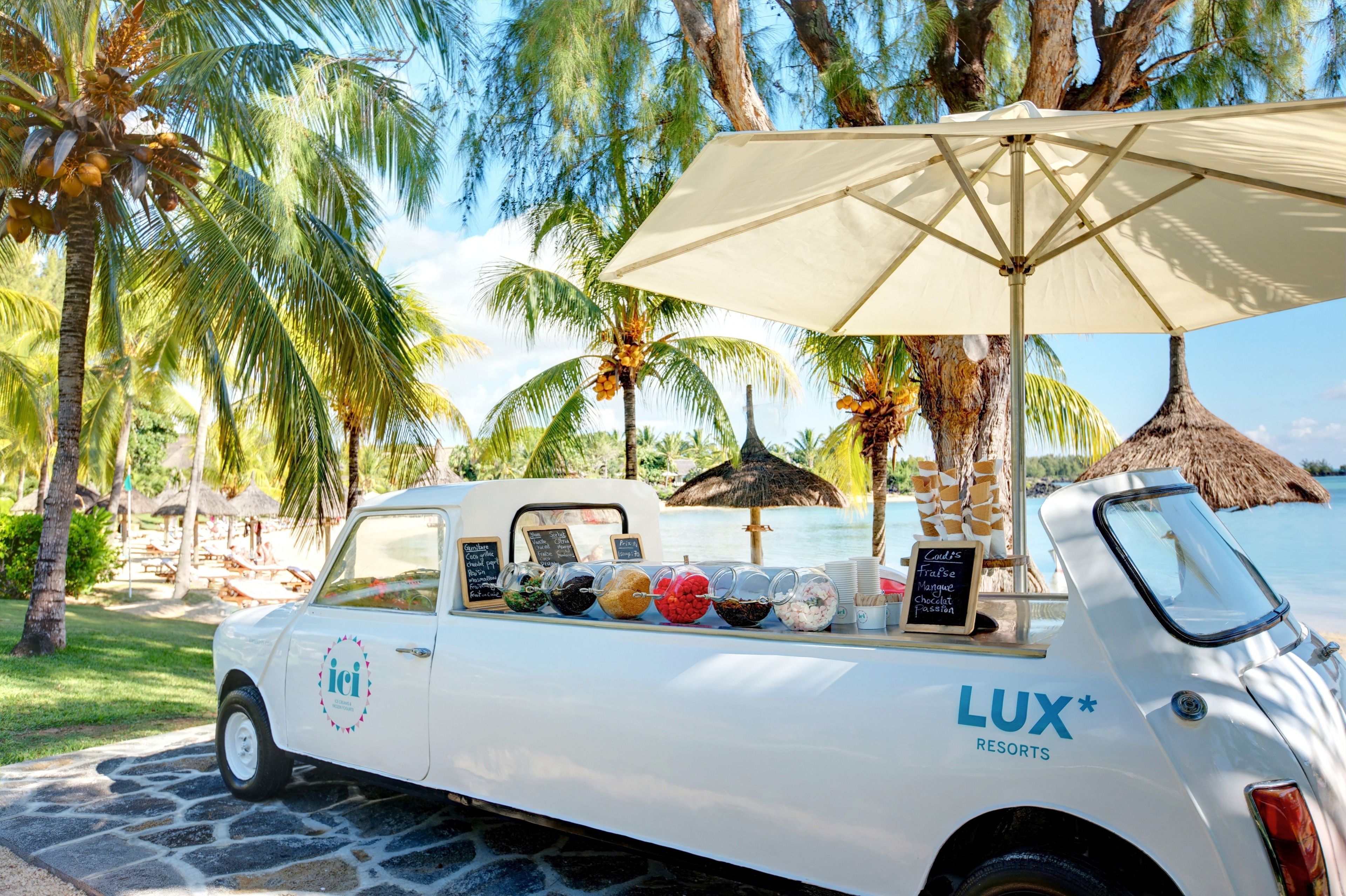 A white food truck with a canopy, branded with "LUX* RESORTS," parked on a stone pathway with palm trees and a beach in the background.