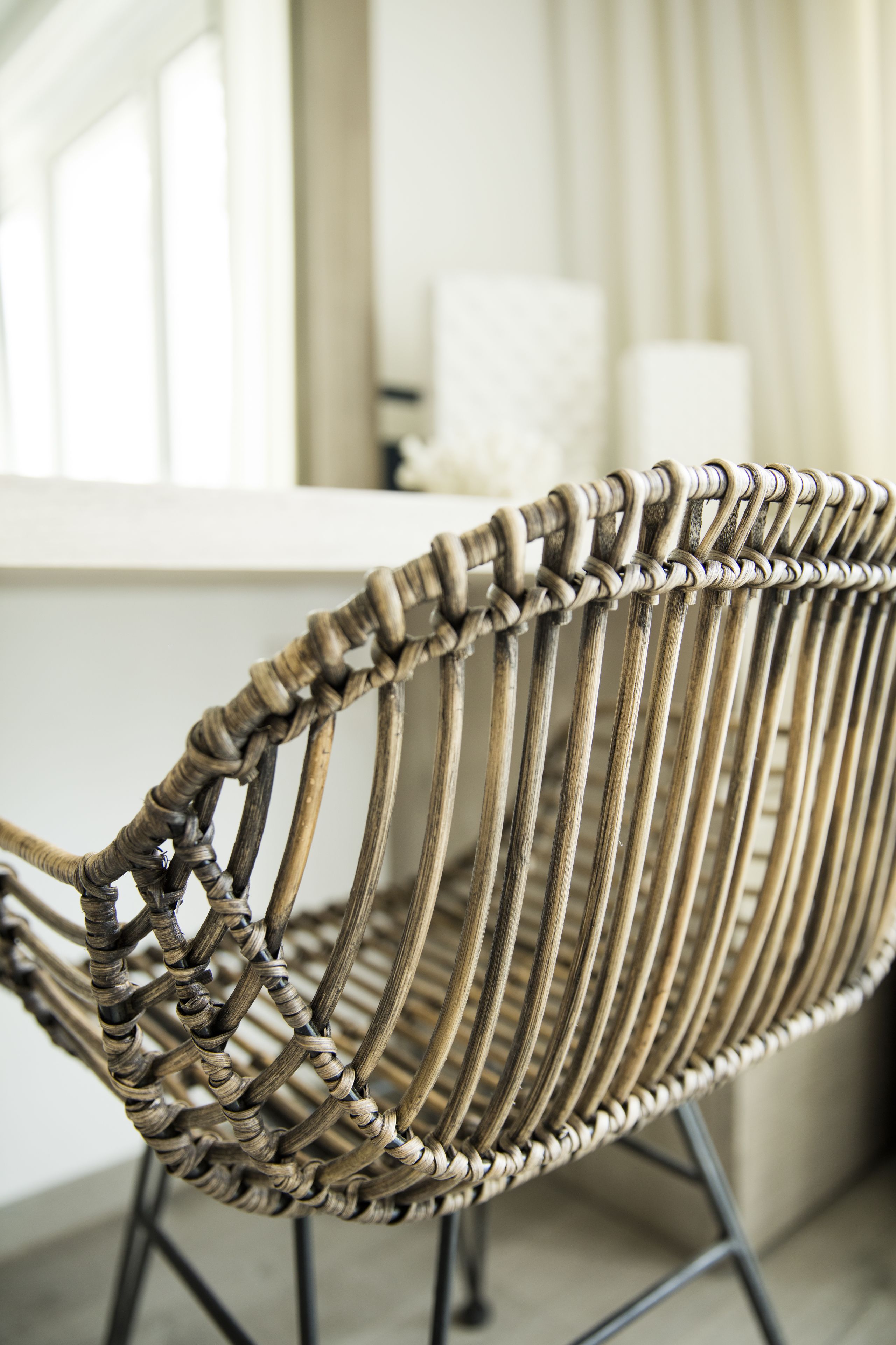 Una silla de mimbre con respaldo tejido y patas metálicas, ubicada en una habitación con un mostrador blanco y una ventana al fondo.