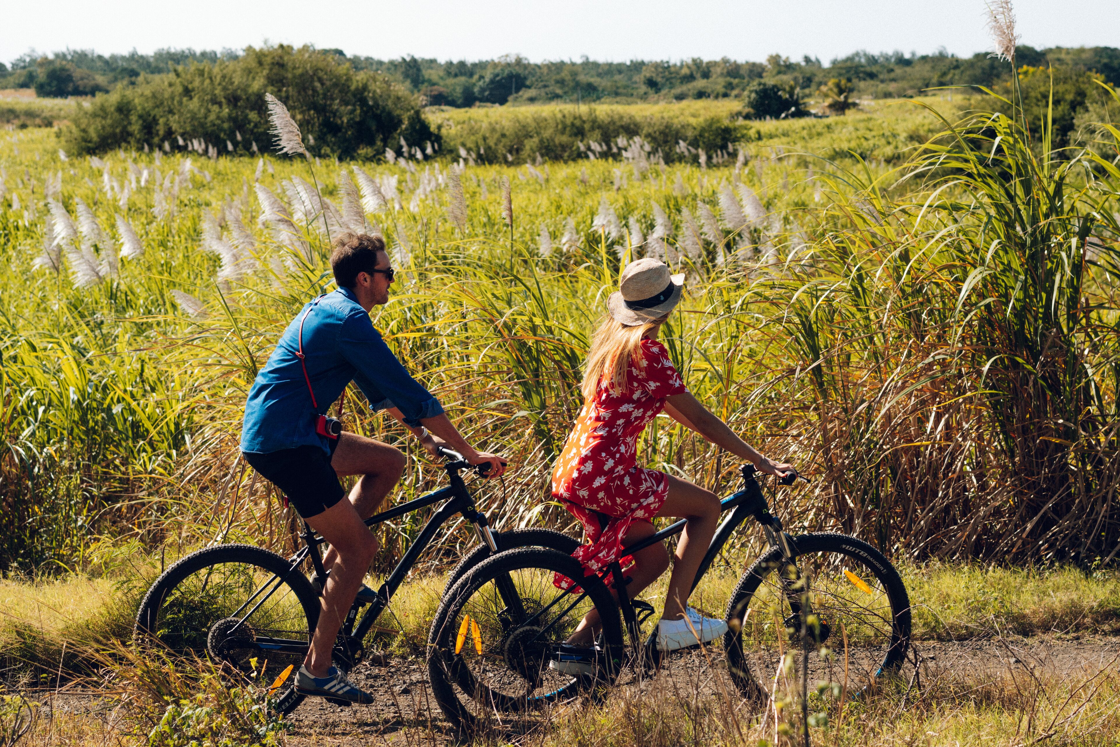 Una pareja en bicicleta en un campo con hierba alta y árboles al fondo.