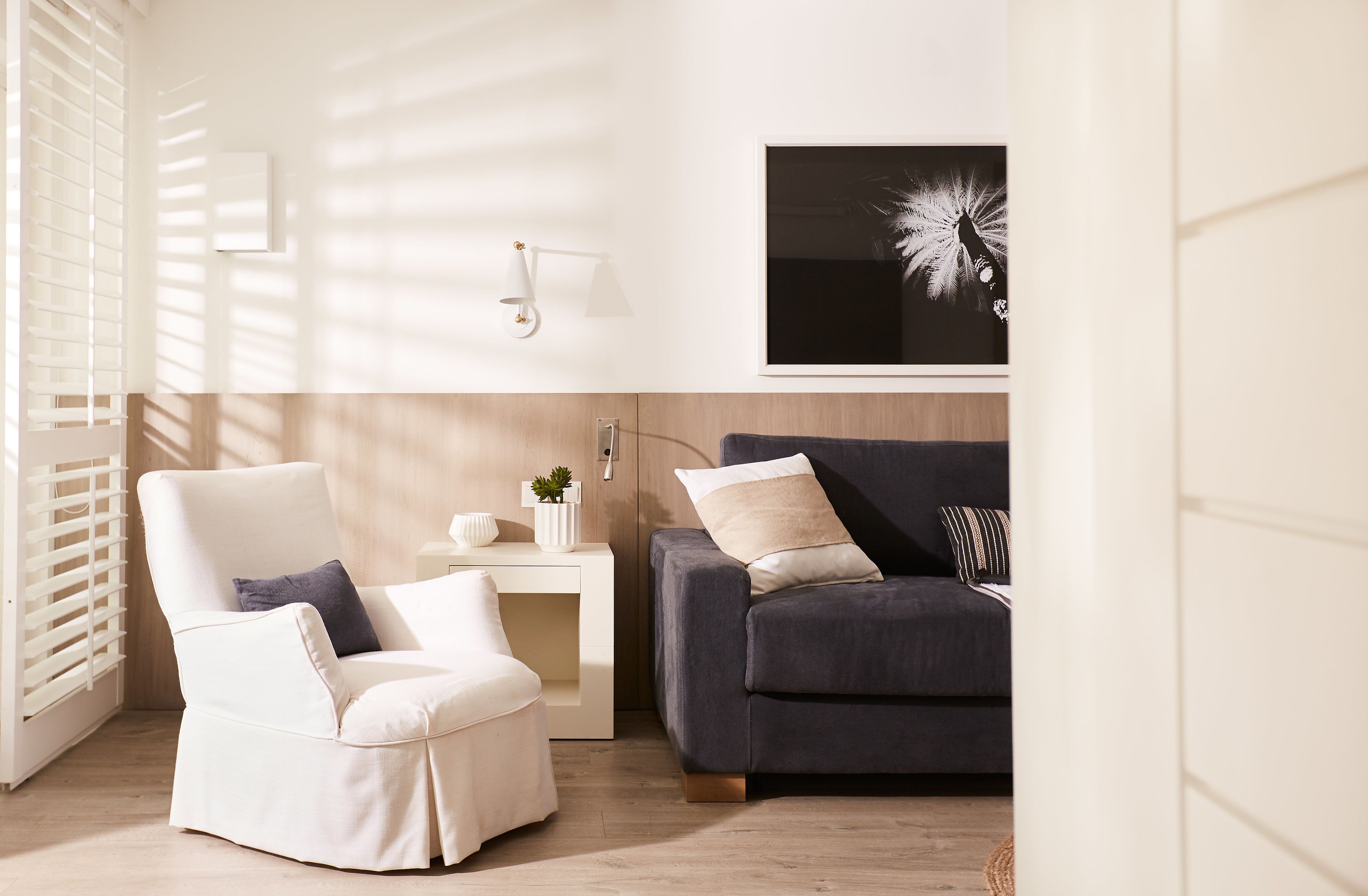 A modern living room with a white armchair, a dark sofa, a small side table with a plant, and a wall-mounted lamp.