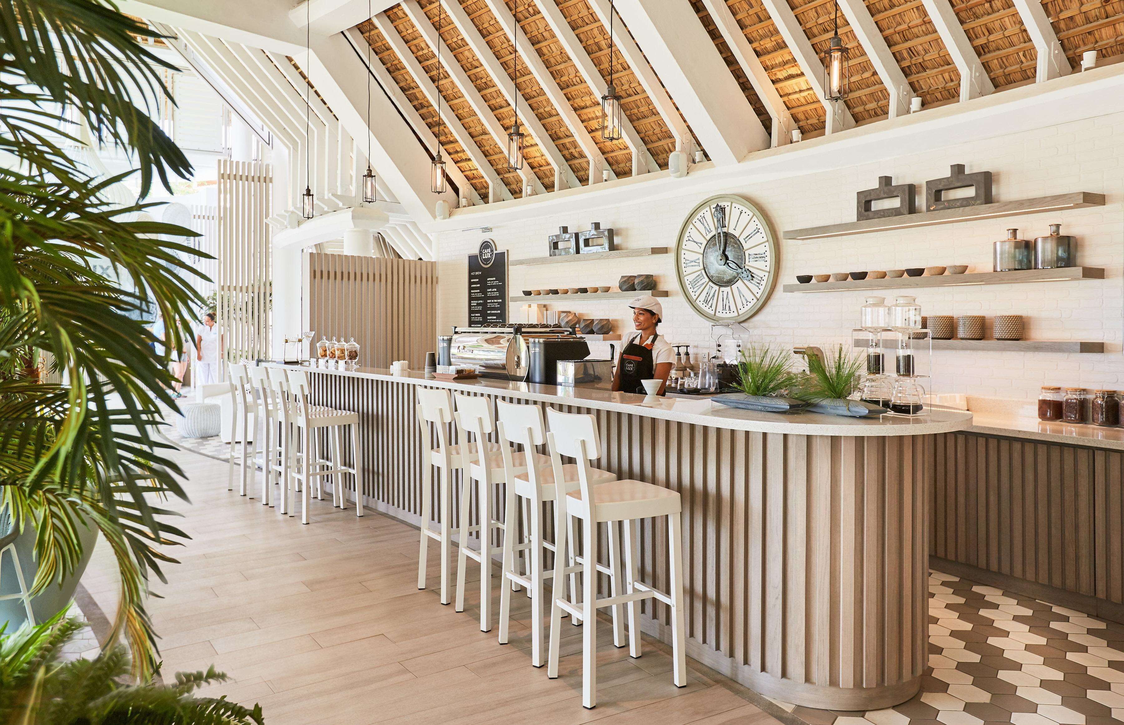A modern bar area with a high ceiling, wooden beams, and a large clock on the wall. There are bar stools and a counter with a coffee machine.