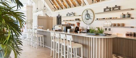 A modern bar area with a high ceiling, wooden beams, and a large clock on the wall. There are bar stools and a counter with a coffee machine.
