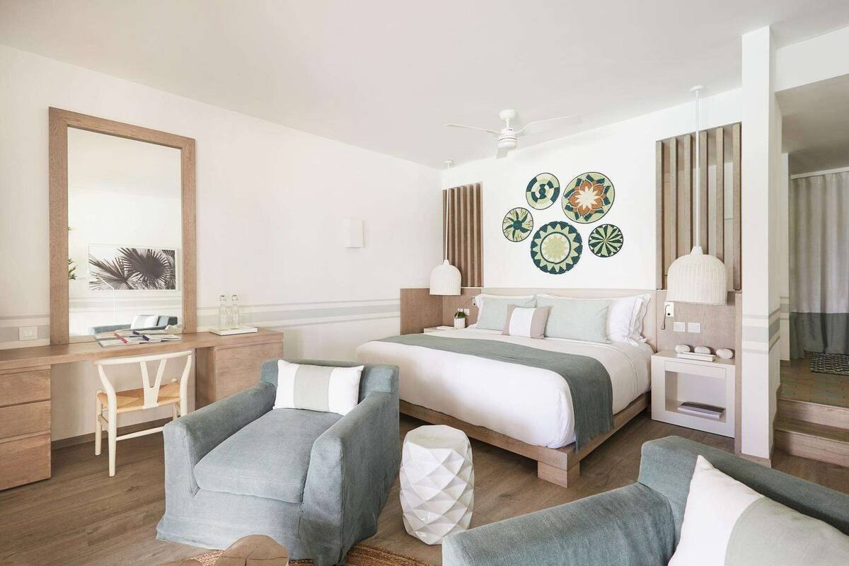 A modern hotel room with a bed, two armchairs, a desk, and a large mirror.