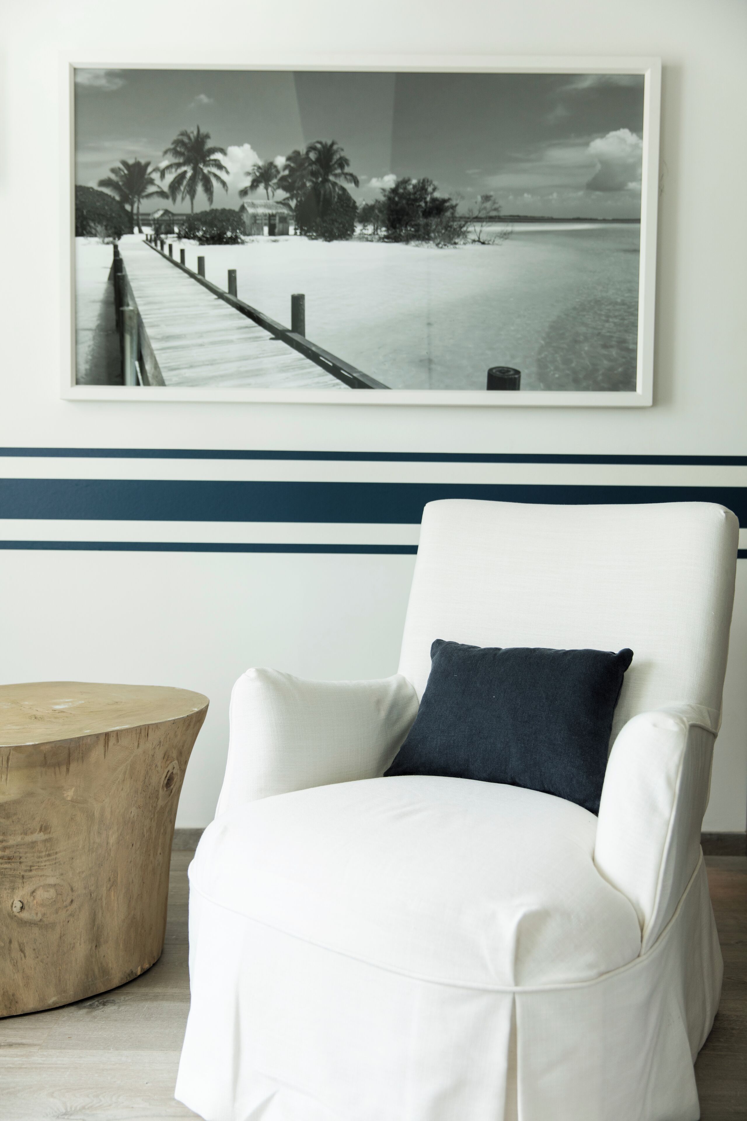 A white armchair with a dark cushion, a wooden side table, and a framed black and white photograph of a beach scene on the wall.