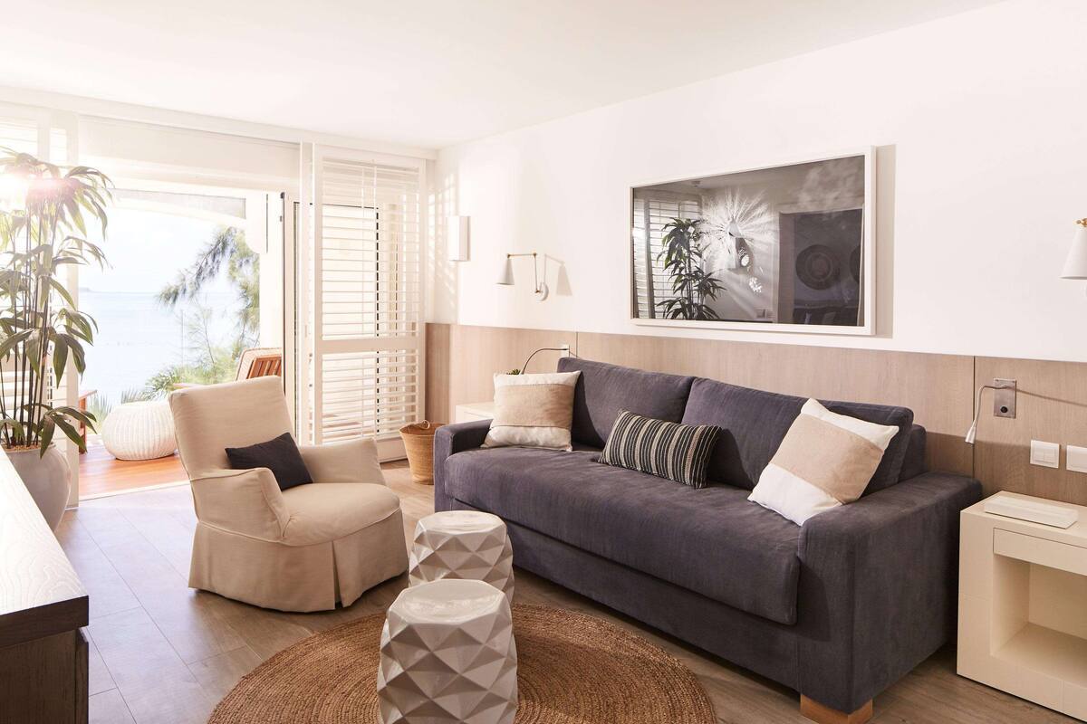 A modern living room with a grey sofa, beige armchairs, and a large window with a view of the ocean.