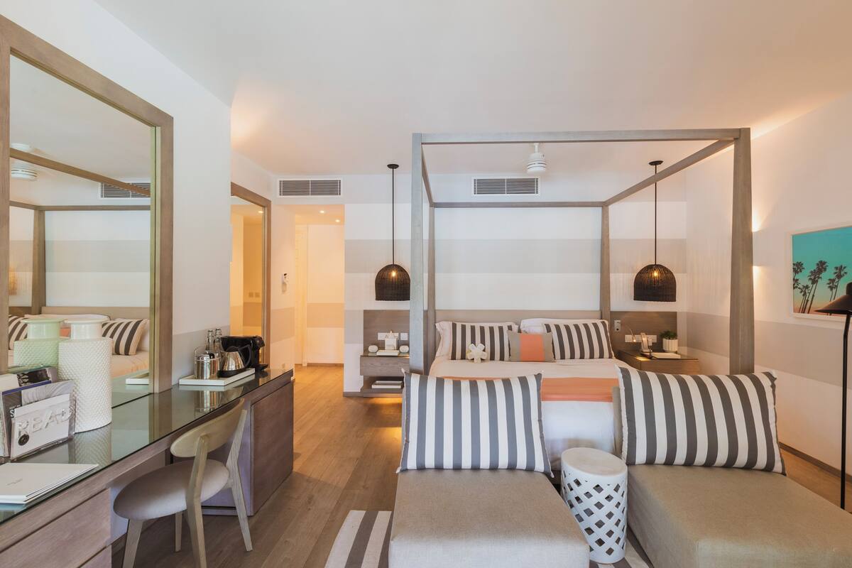 A modern hotel room with a four-poster bed, a desk with a chair, and a large mirror.