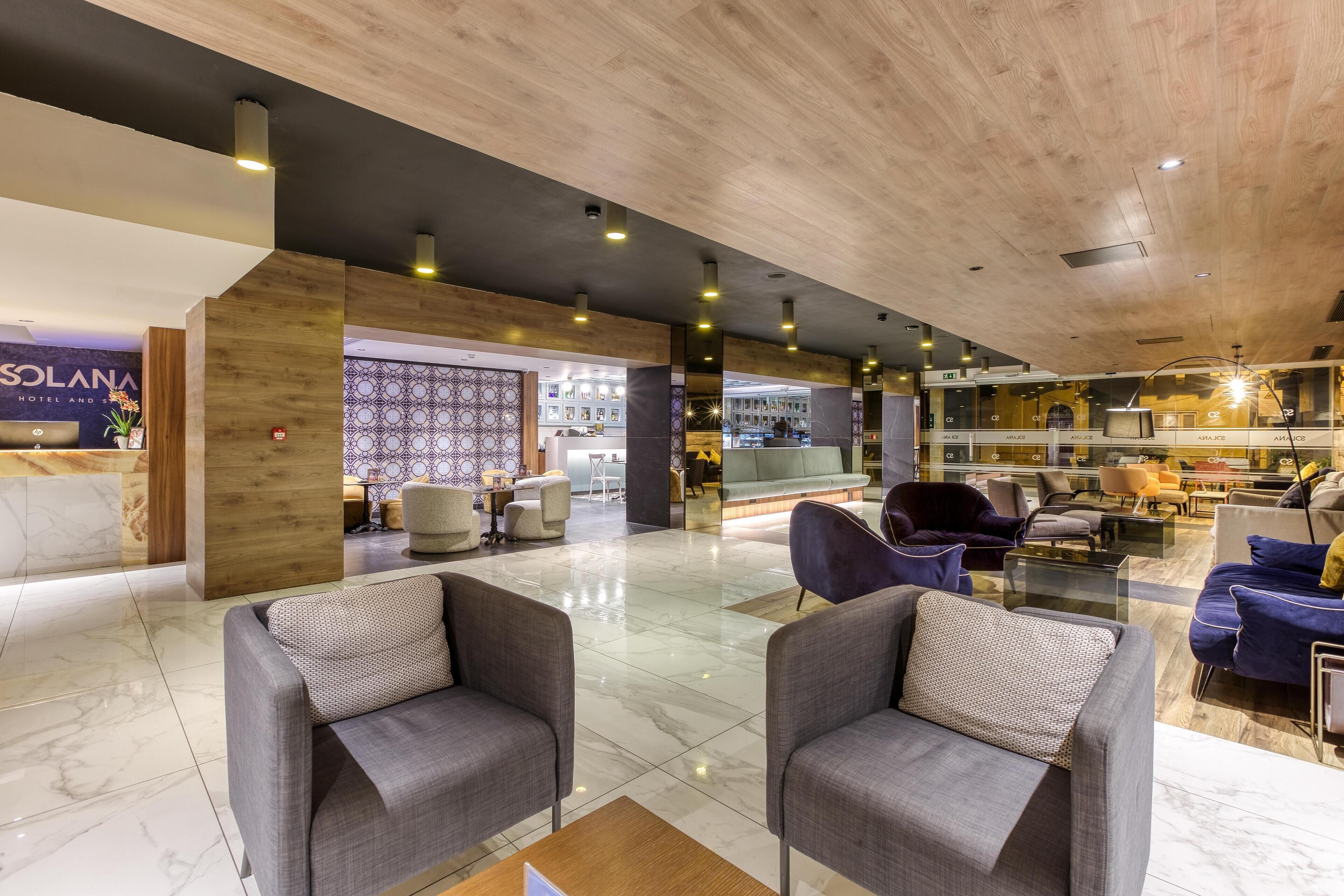 Un moderno vestíbulo de hotel con techo de madera, suelo de mármol y una zona de estar con sofás y sillones grises.