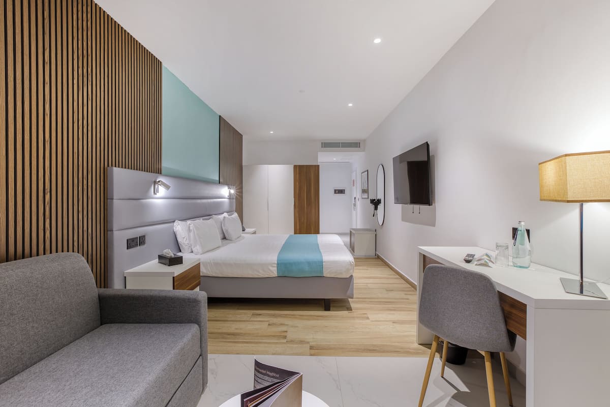 Quarto de hotel moderno com cama, escrivaninha, poltrona, sofá e televisão.
