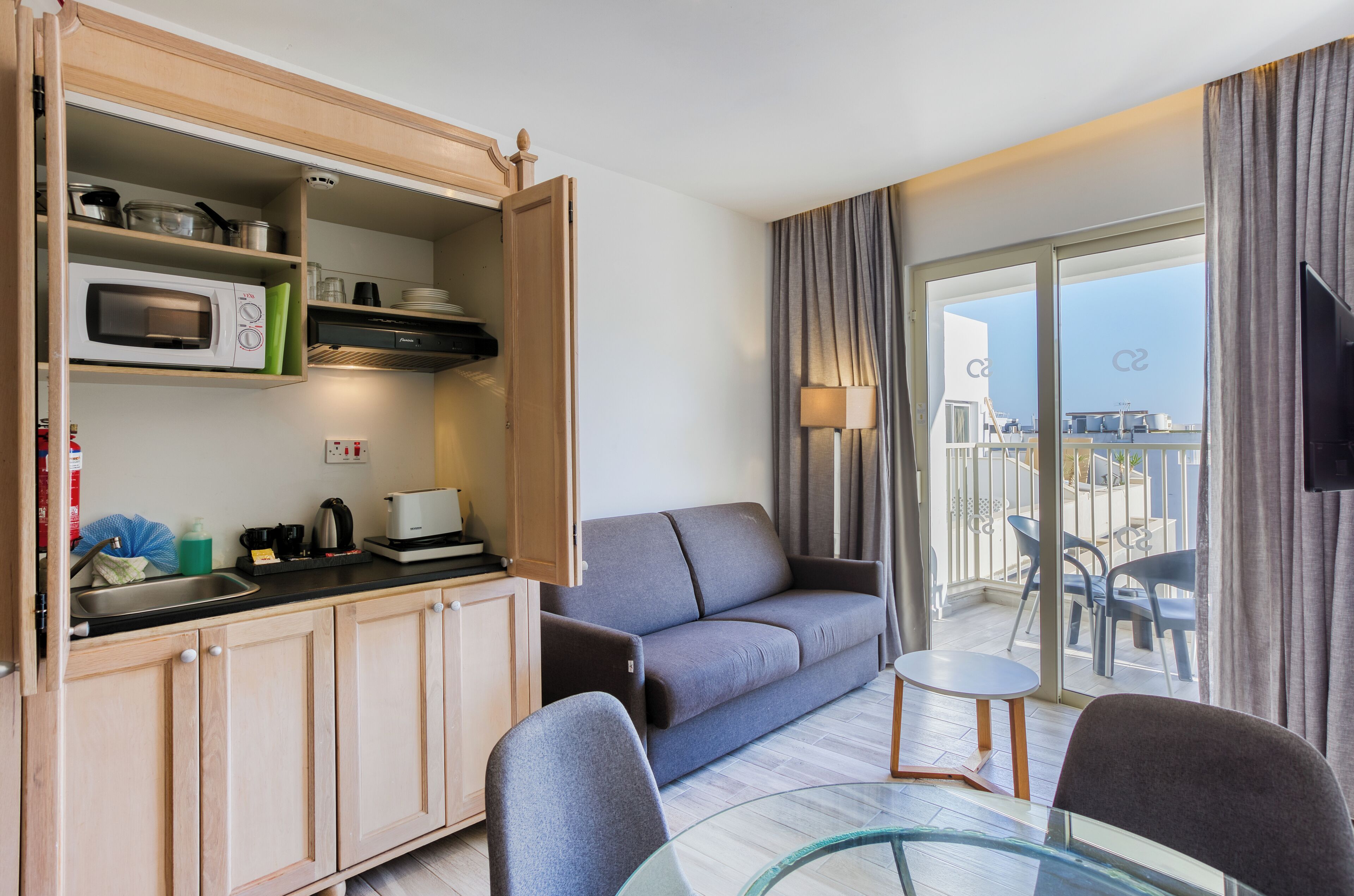 A compact living space with a kitchenette, a sofa, a glass table, and a balcony with a view.